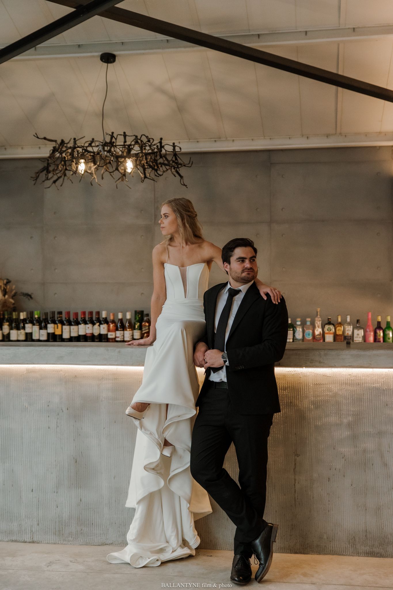 Bride in white ruffled gown stands elevated beside groom in black suit at elegant bar venue with bottle display.