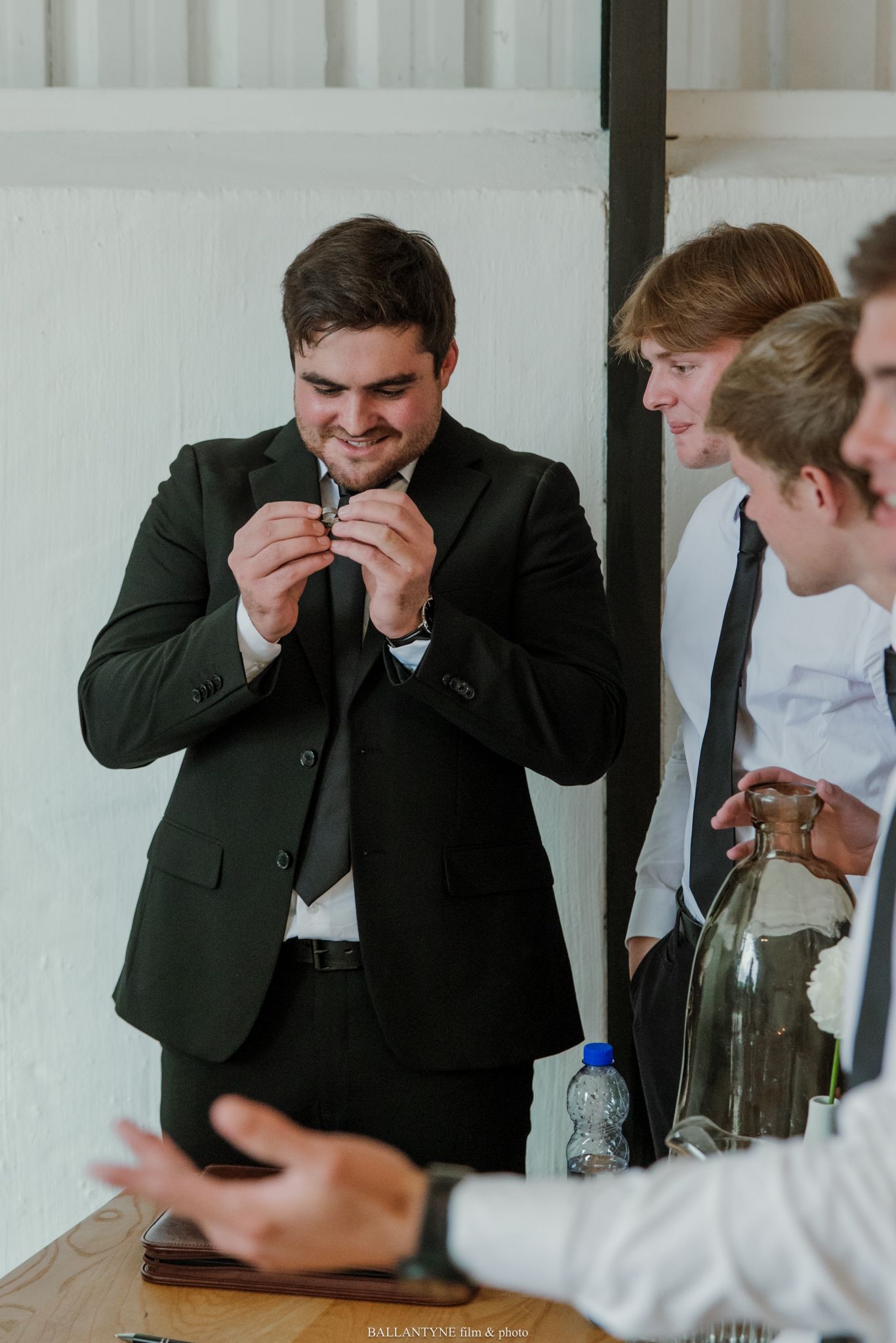 Groom in black suit adjusting tie while smiling, surrounded by groomsmen in white shirts at wedding preparation.