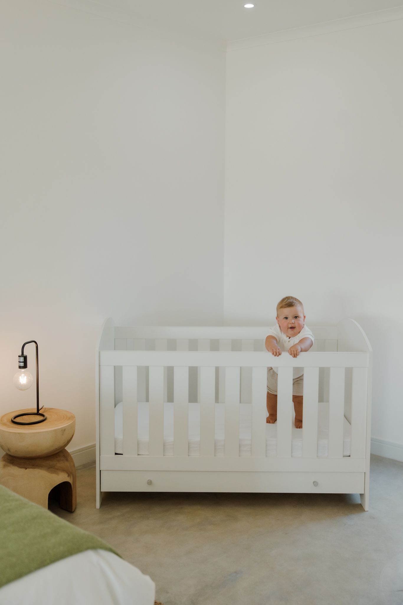 A baby in white clothing stands and smiles inside a modern white crib in a minimalist nursery room.