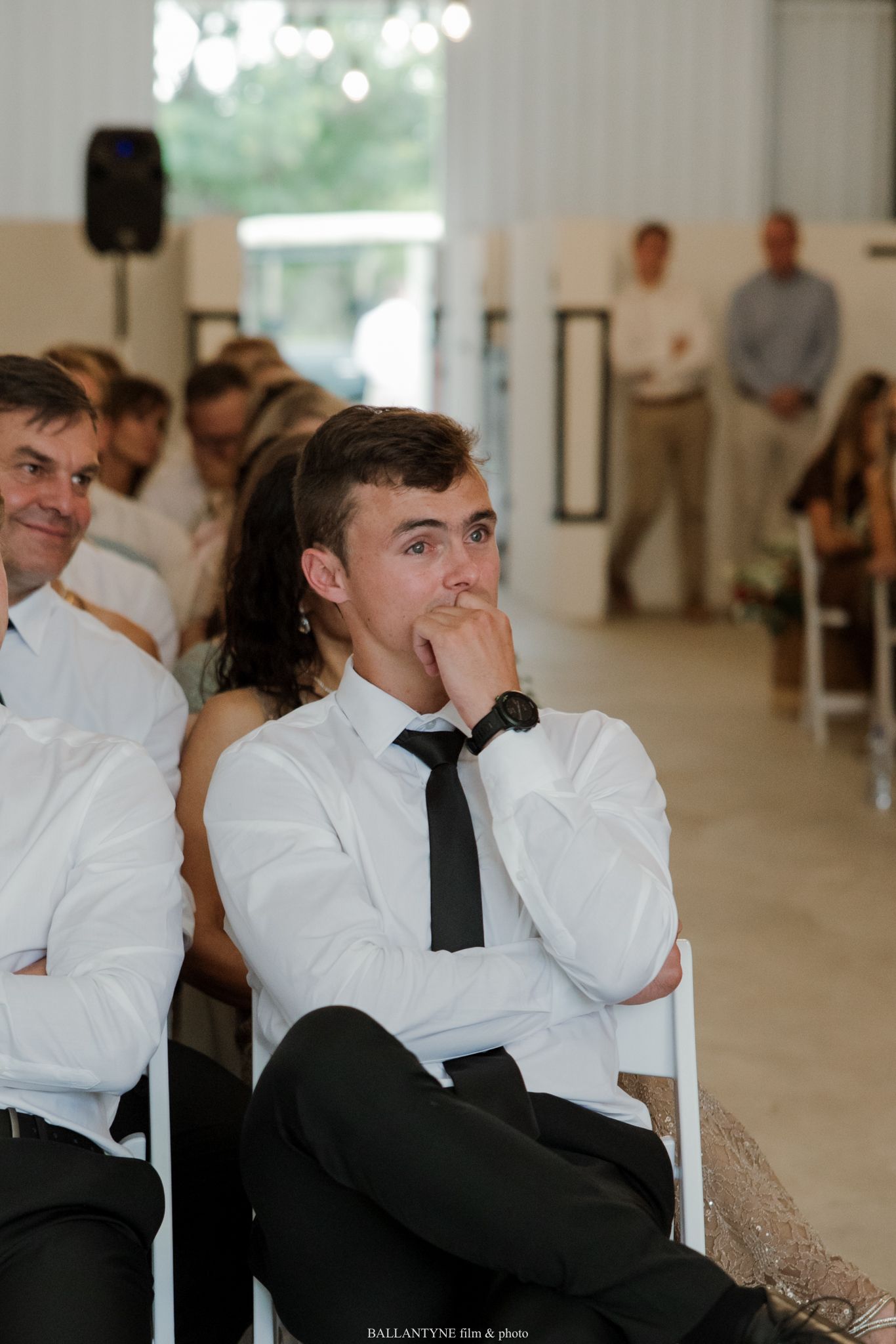 Young man in white shirt and black tie sits thoughtfully at a wedding ceremony, hand raised to his mouth.