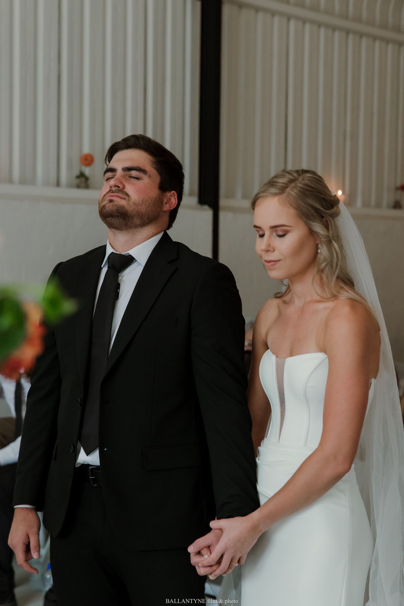 Bride in strapless white gown and veil holds hands with groom in black suit during emotional wedding ceremony.