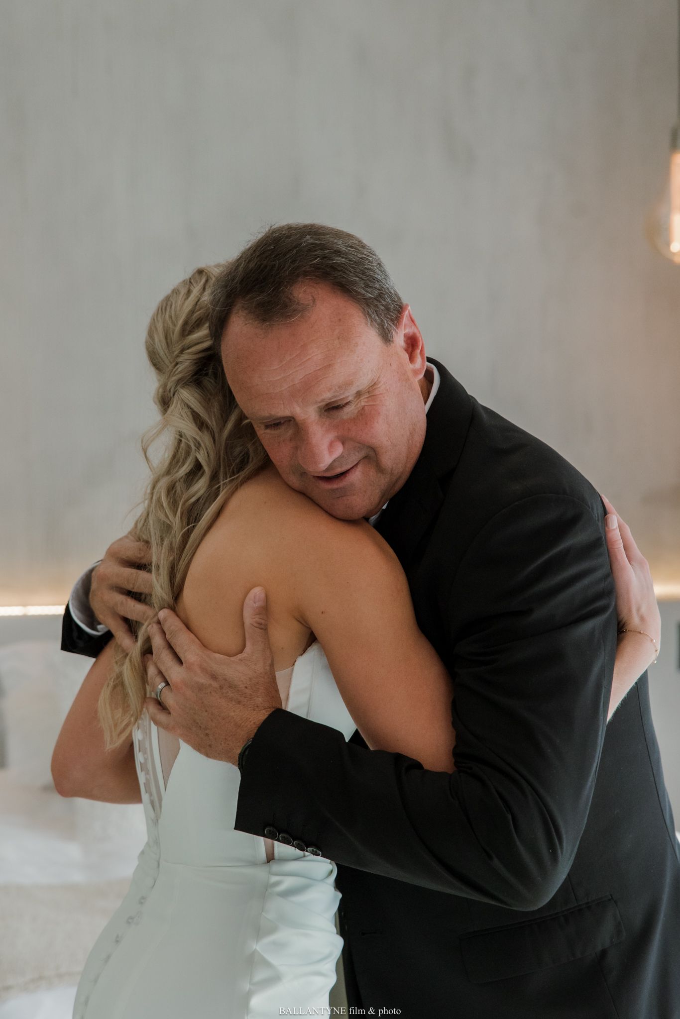 A bride in a white gown shares an emotional hug with an older man in a black suit during a wedding reception.