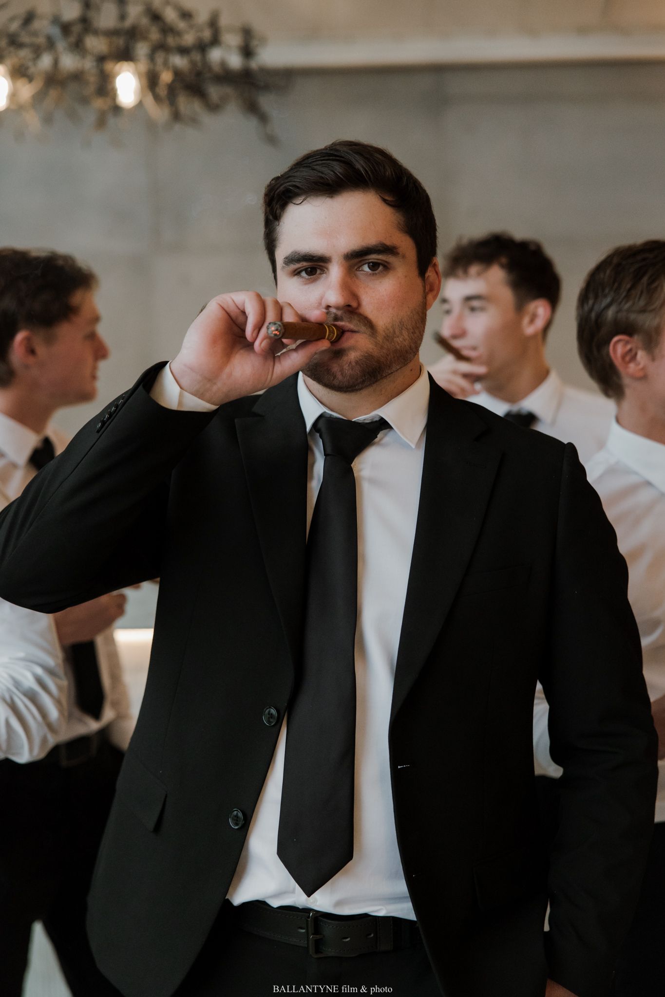 A man in a black suit and tie smoking a cigar at a formal event with guests in the background.