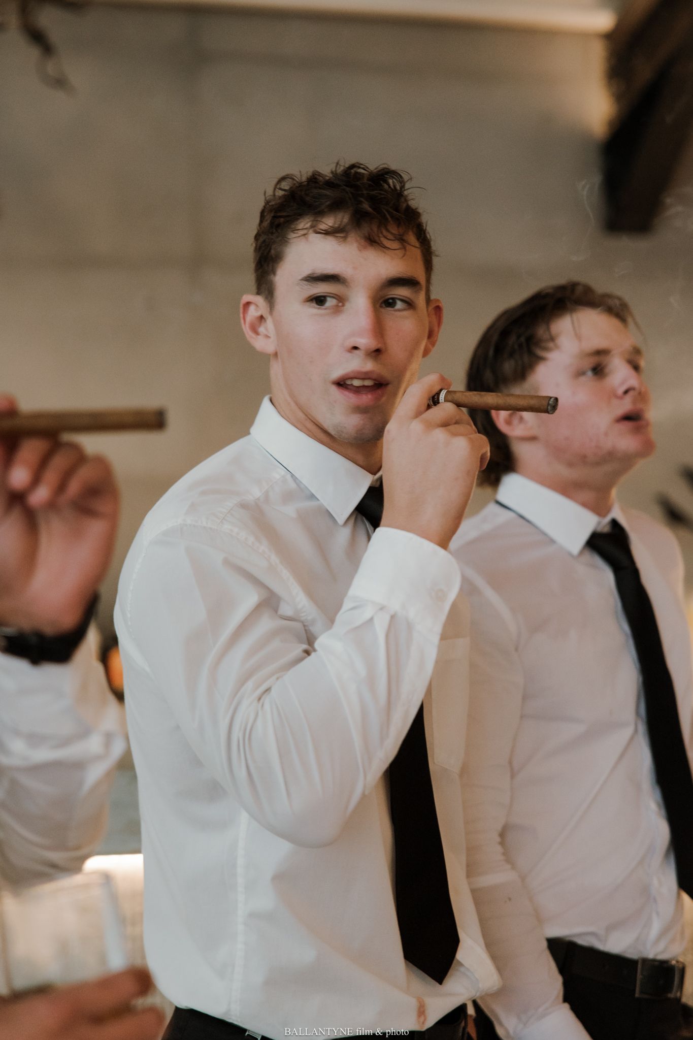 Young men in white dress shirts and black ties smoking cigars at a formal event or wedding celebration.