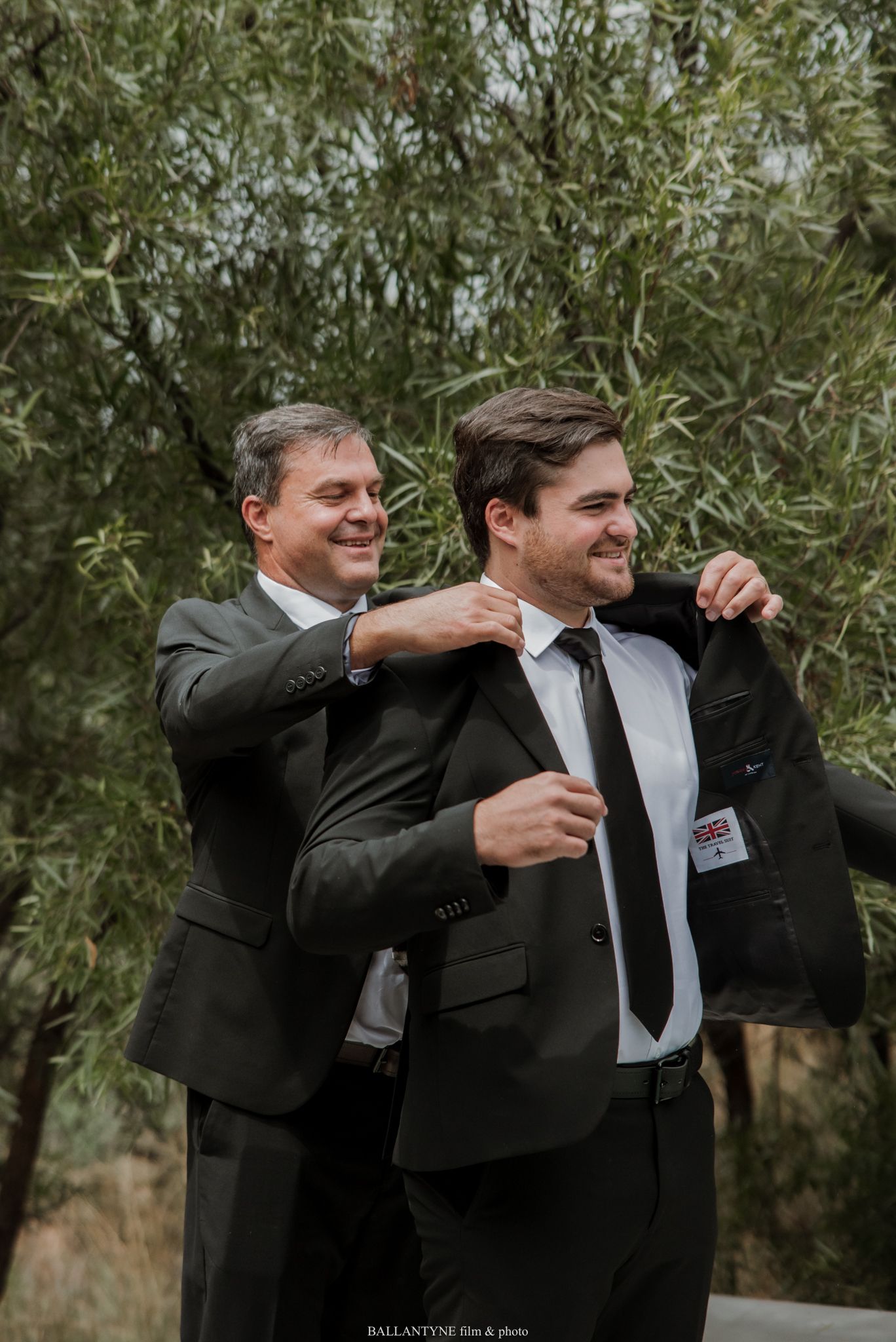 Man helping groom adjust suit jacket outdoors before wedding ceremony.