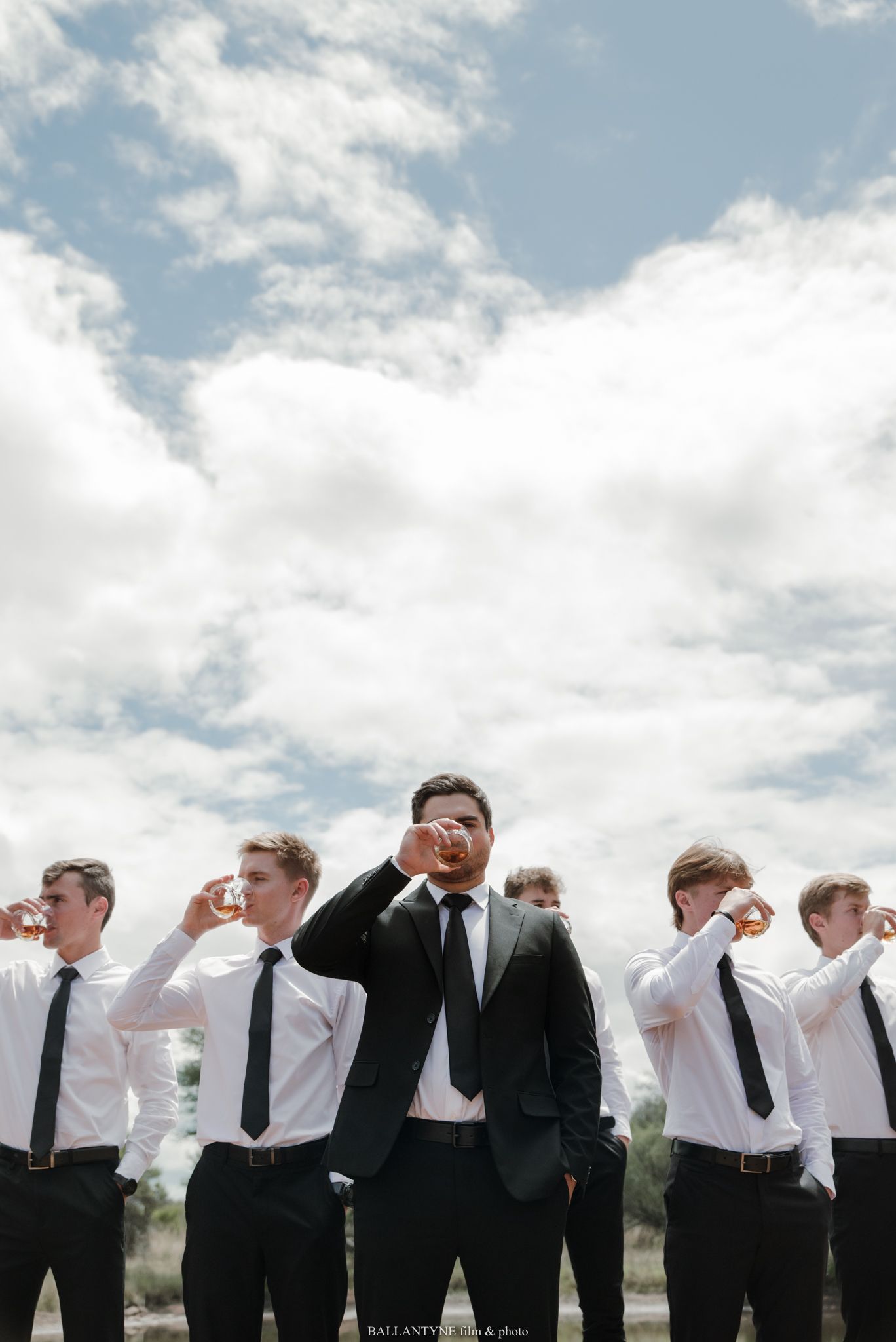 Groom in dark suit raises a toast with groomsmen in white shirts and black ties outdoors under a cloudy sky.