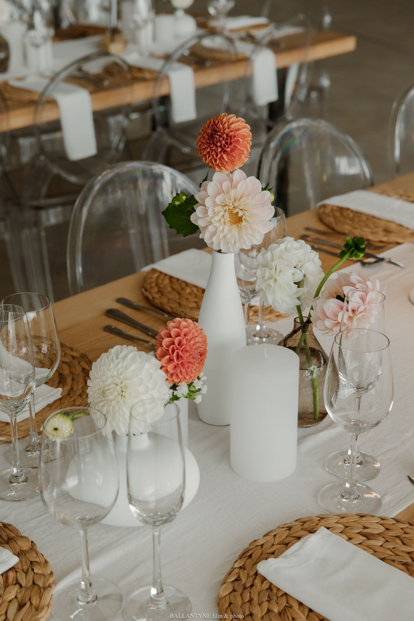 Elegant wedding table with white dahlias, pink blooms, wicker chargers, clear ghost chairs, and white candles.