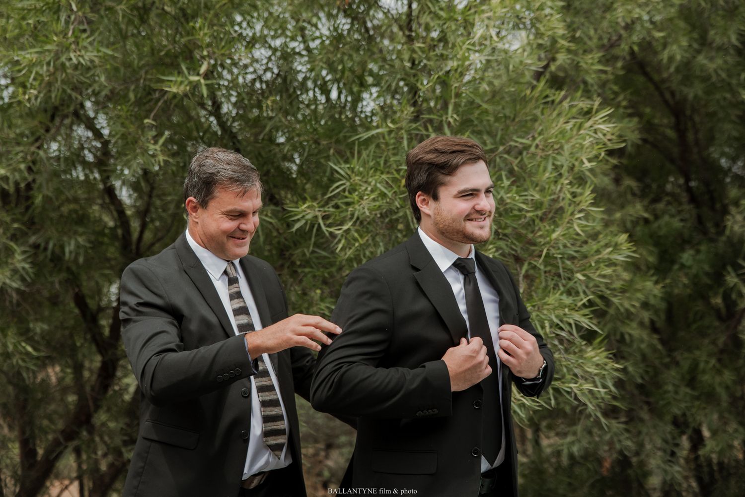 Two men in dark suits adjusting their ties outdoors among green trees, likely at a wedding ceremony.