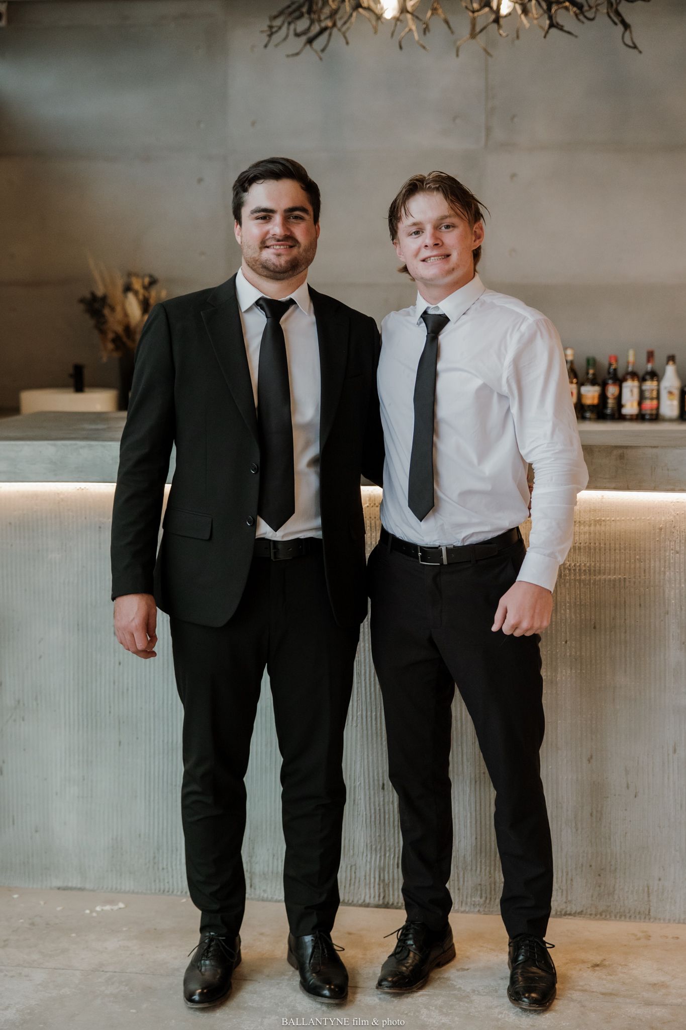 Two young men in black ties and dress shirts smile together in front of a modern bar backdrop.