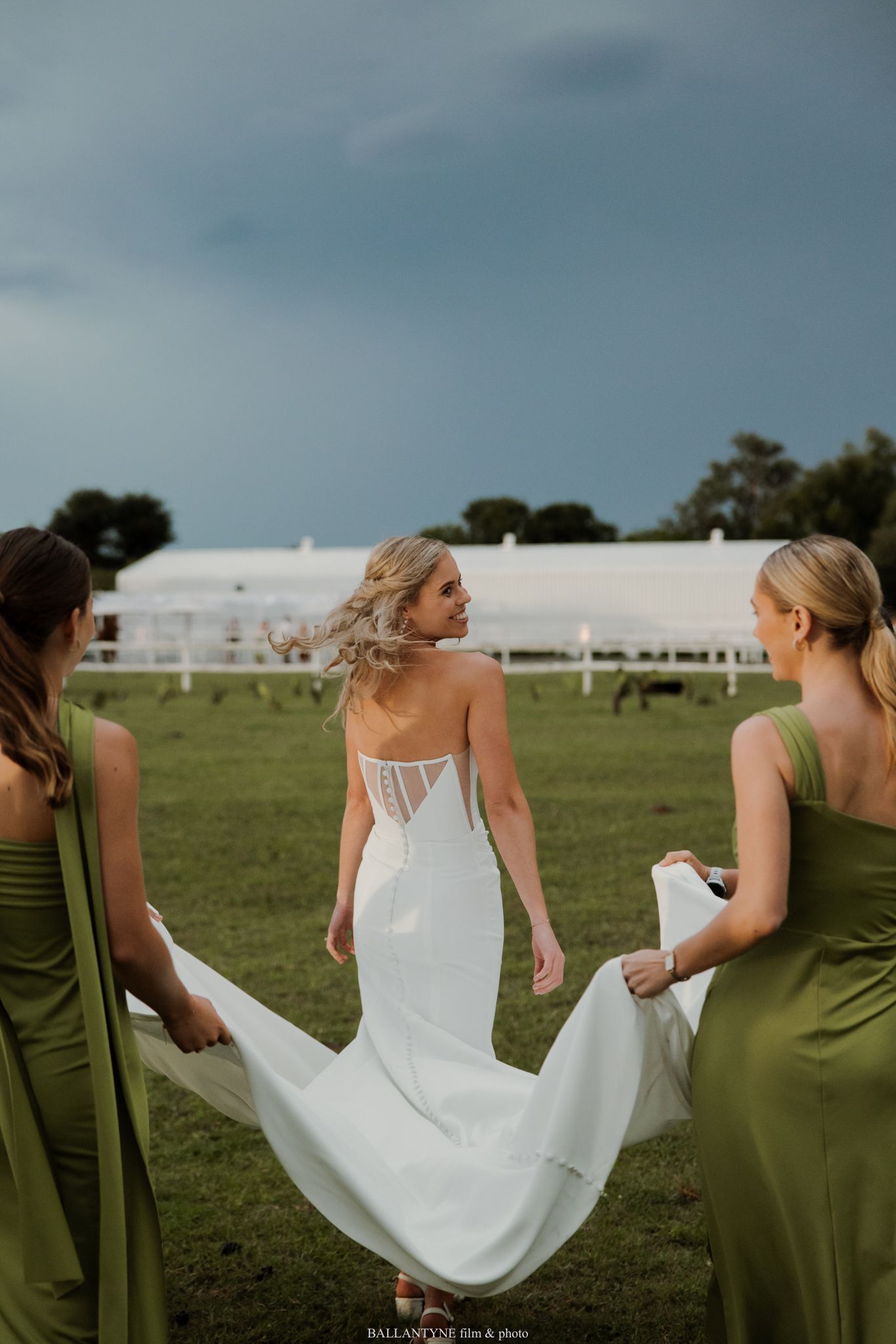 Bride in white backless gown walks through green field as bridesmaids in olive dresses hold her train.