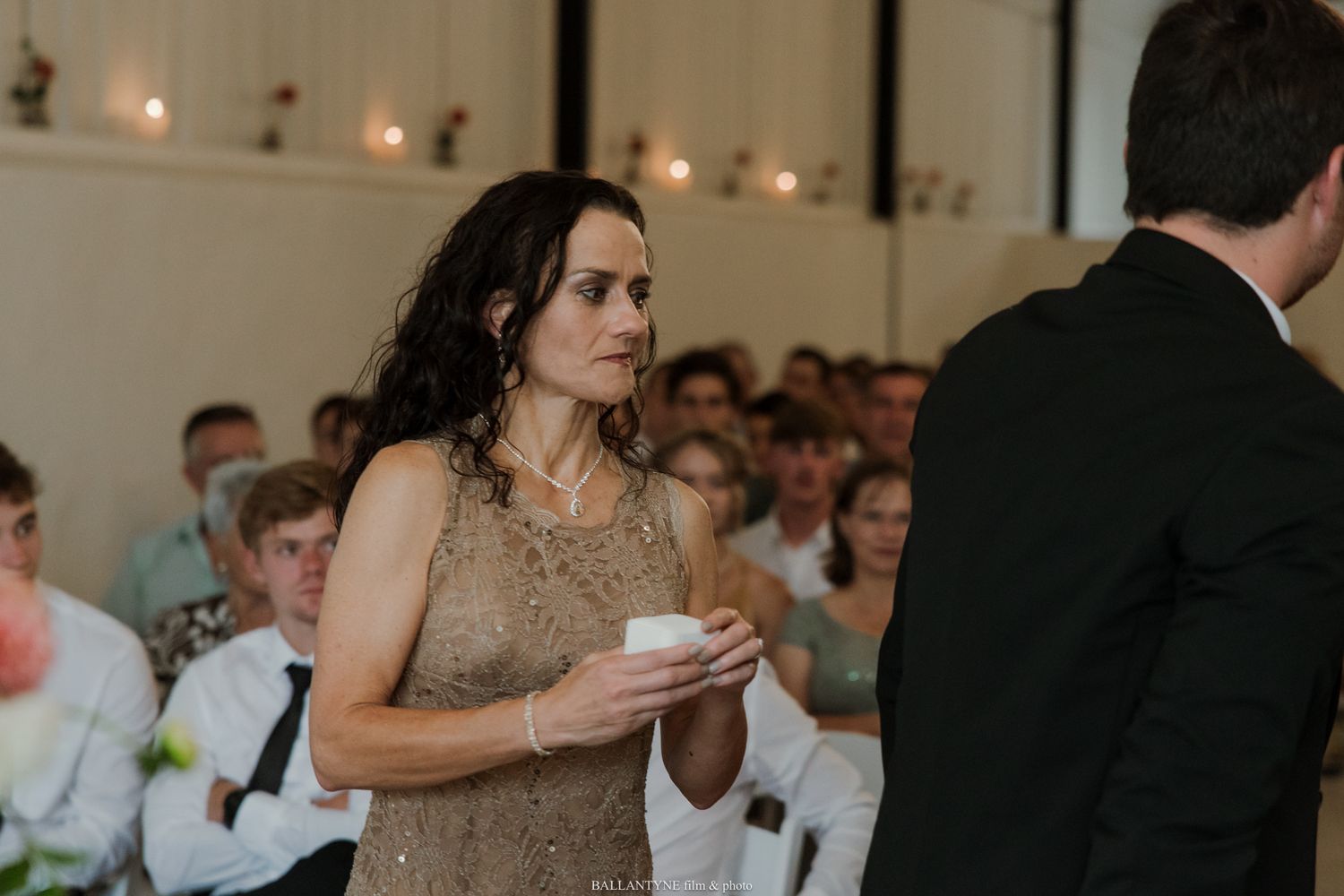 Woman in gold dress holding a candle at an elegant wedding ceremony with guests seated in background.