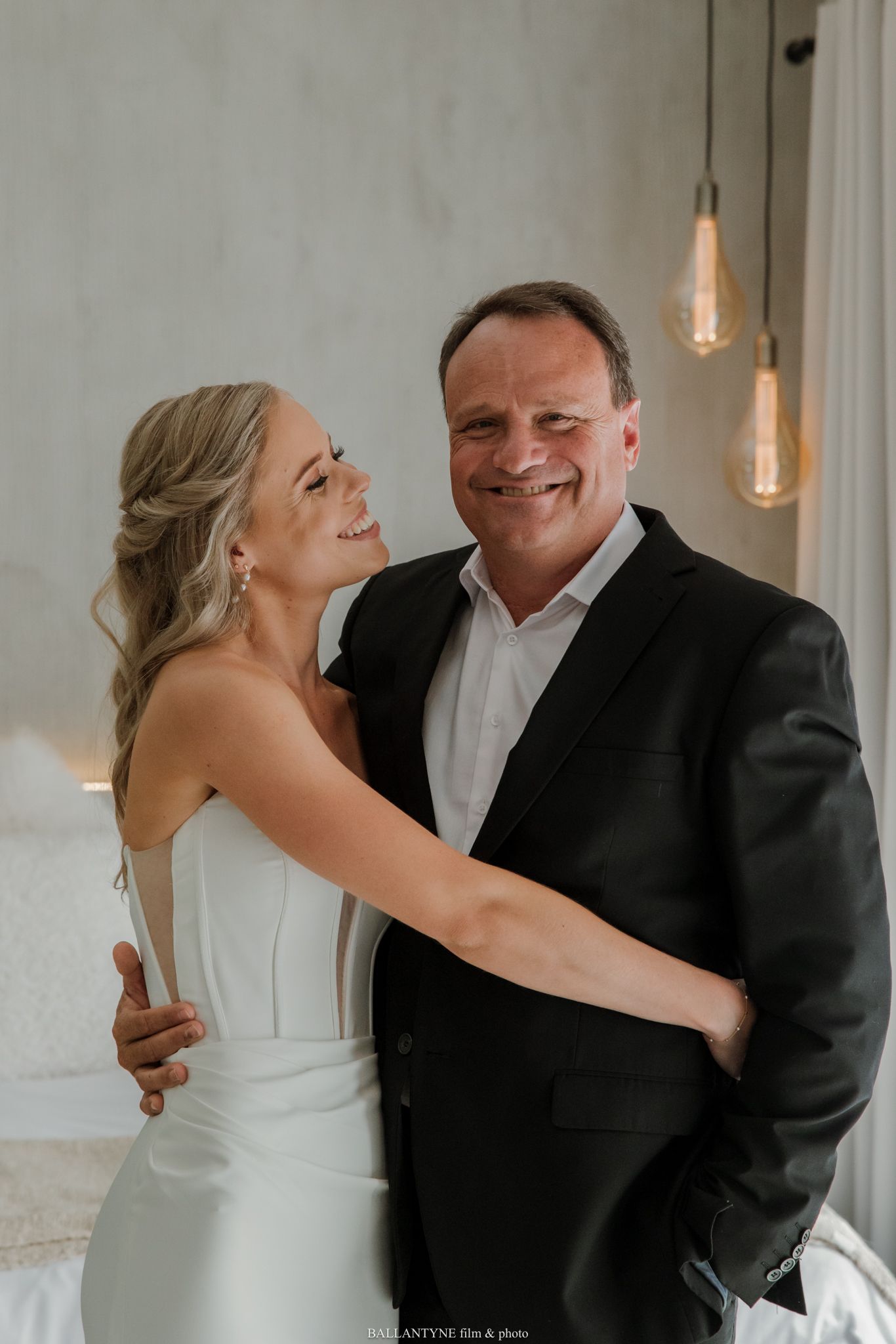 A bride in a white dress embraces a smiling man in a dark suit, posing together in a warmly lit elegant room.
