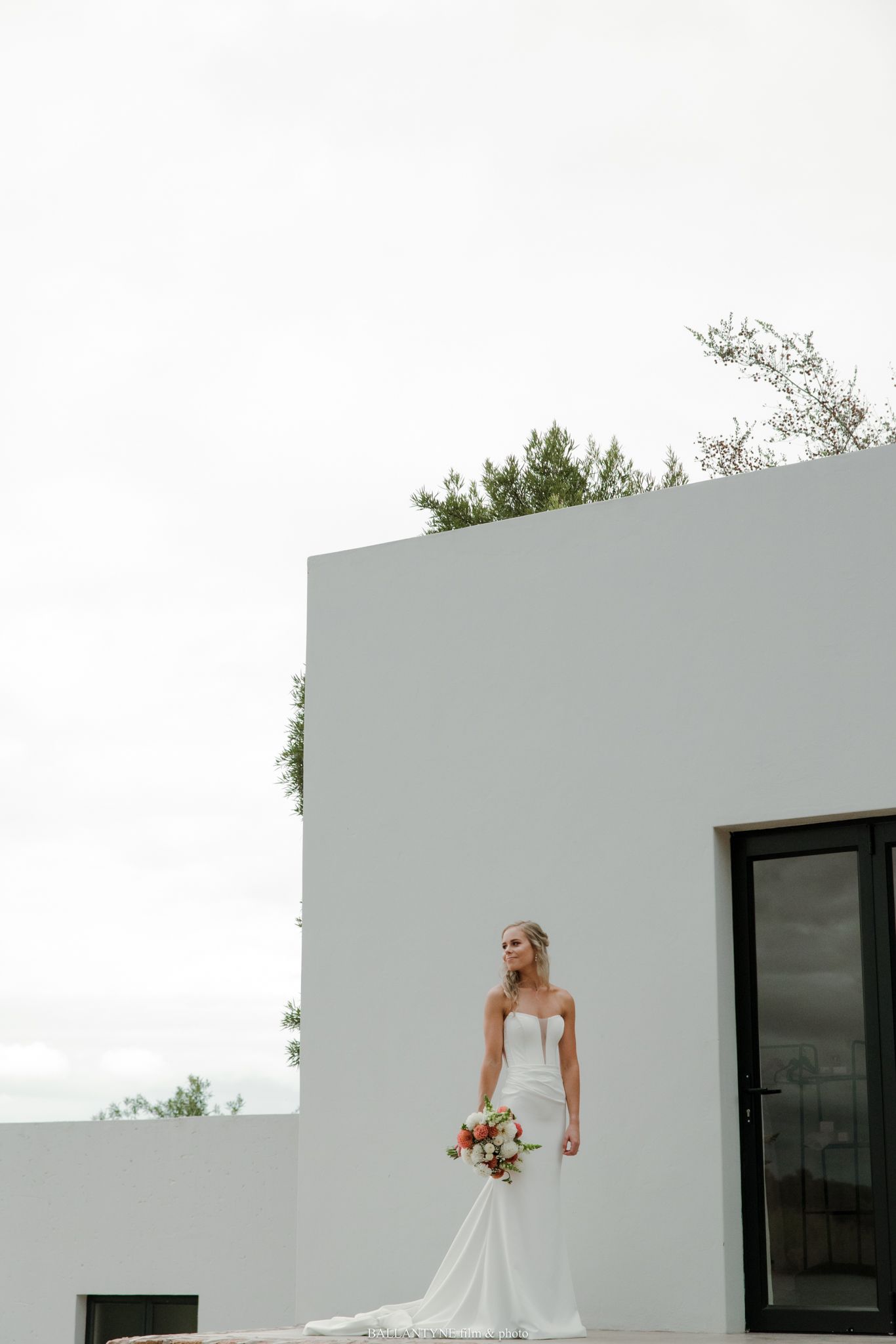 Bride in strapless white gown holding bouquet stands before modern white building with greenery overhead.