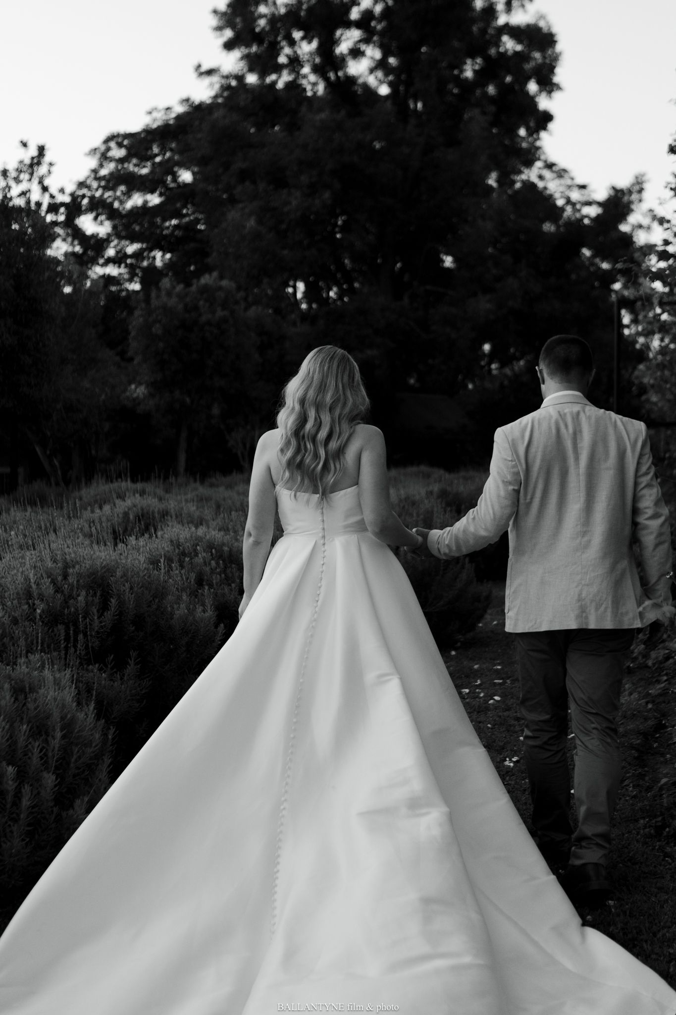 Black and white photo of couple walking, showcasing bride's elegant flowing dress train.