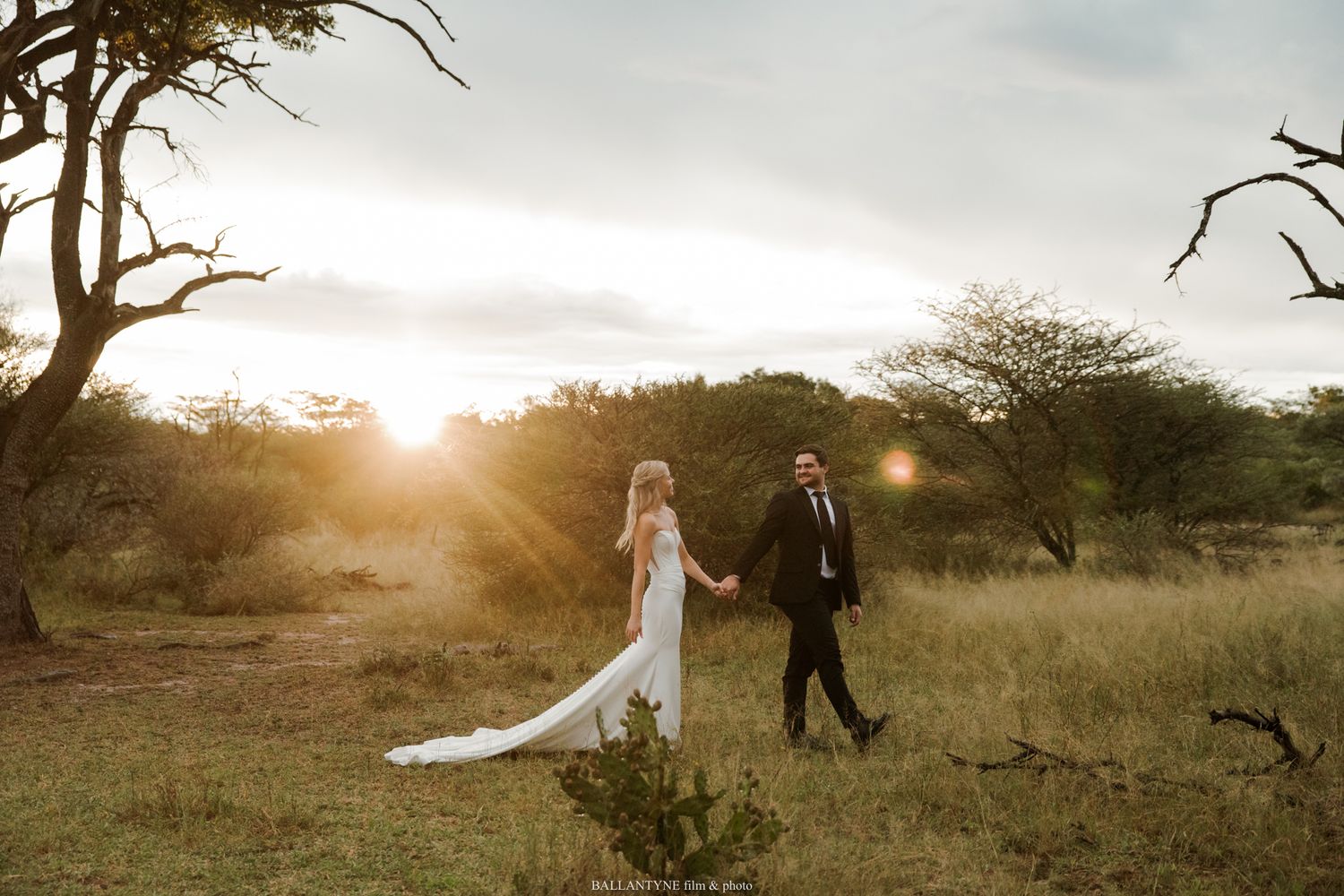 Bride and groom hold hands walking through African savanna at golden sunset, wedding attire, lush greenery.