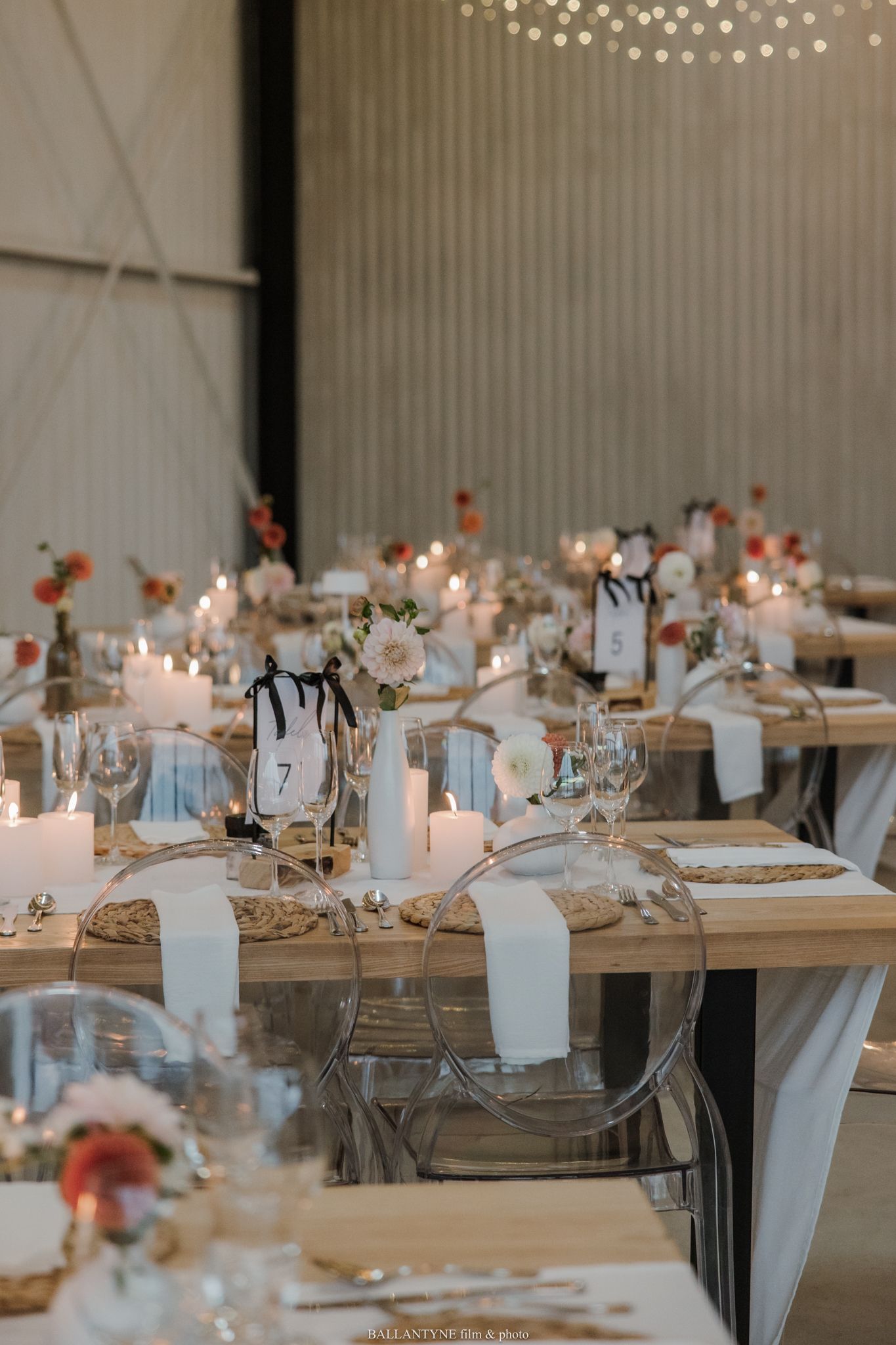 Elegant wedding reception with candles, white napkins, floral centerpieces, and ghost chairs in a rustic barn venue.