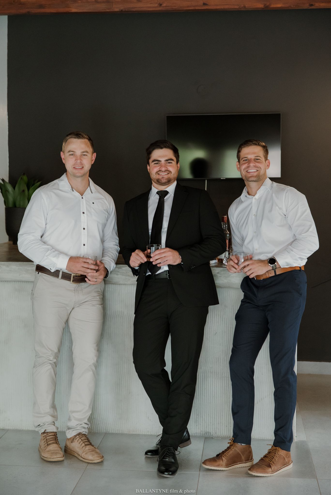 Three men in formal attire smile and hold drinks at an elegant indoor venue.