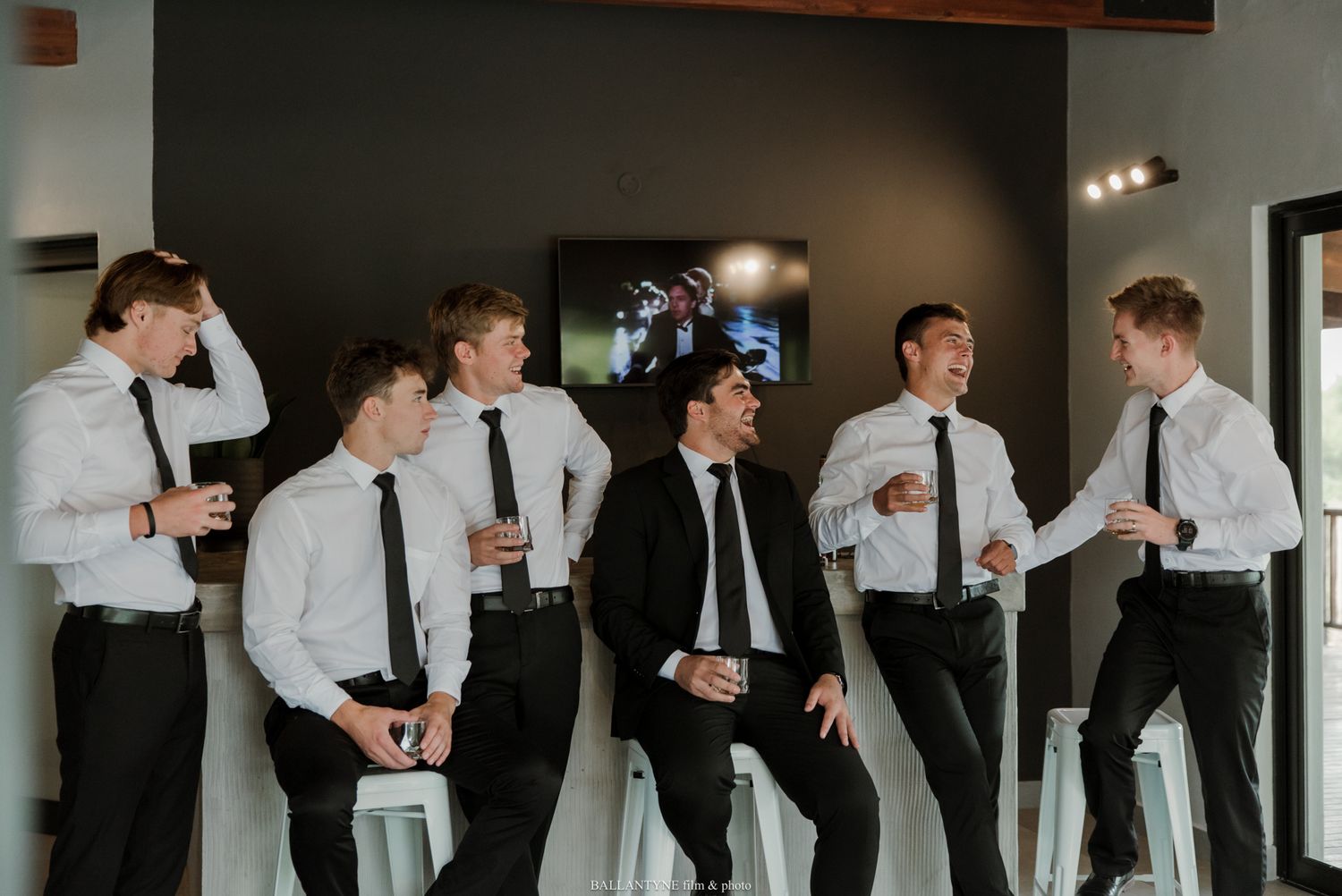 Groomsmen in white shirts and black ties laughing together before a wedding ceremony.