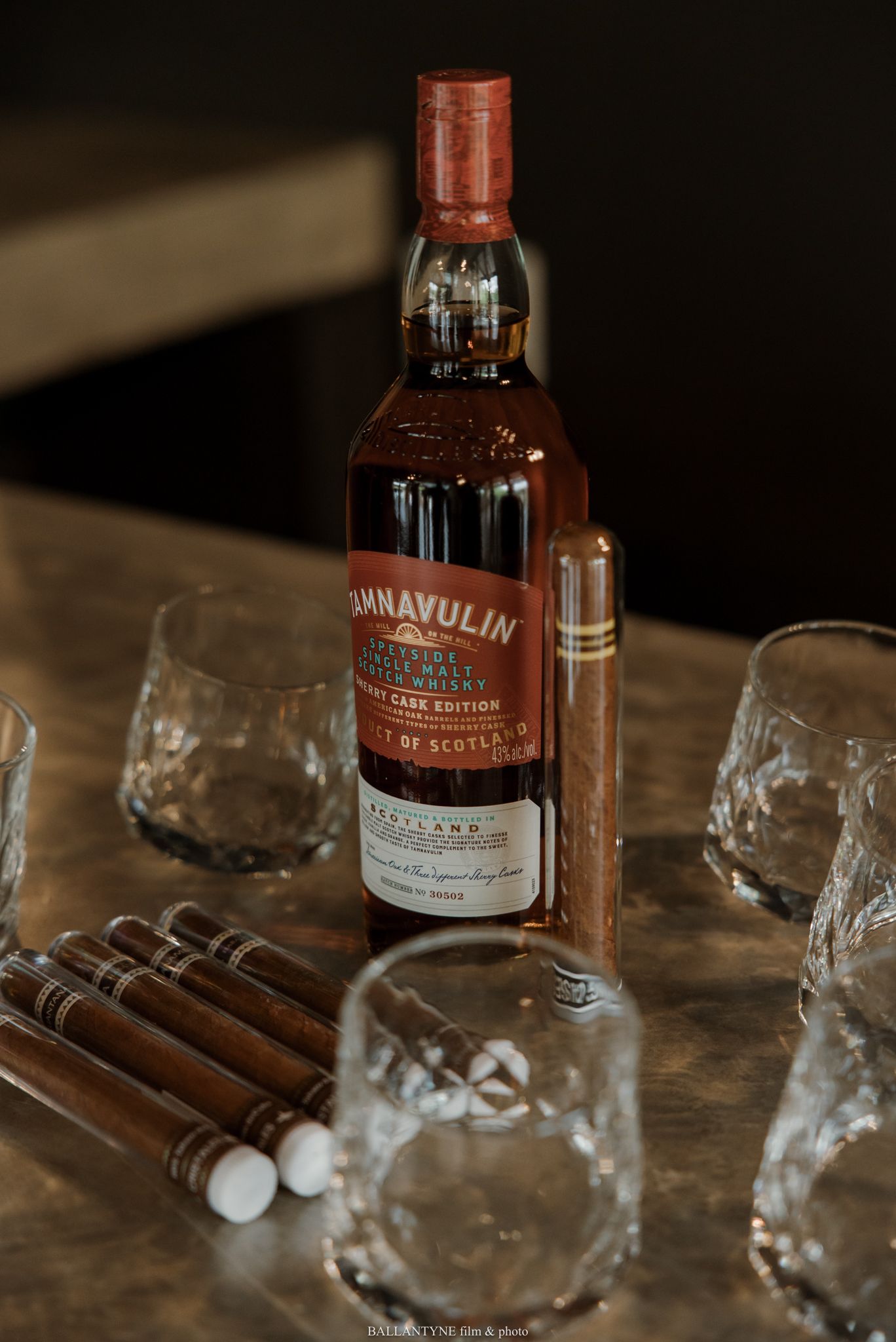 Tamnavulin whisky bottle beside cigars and crystal glasses on a wooden table in a moody, dimly lit setting.