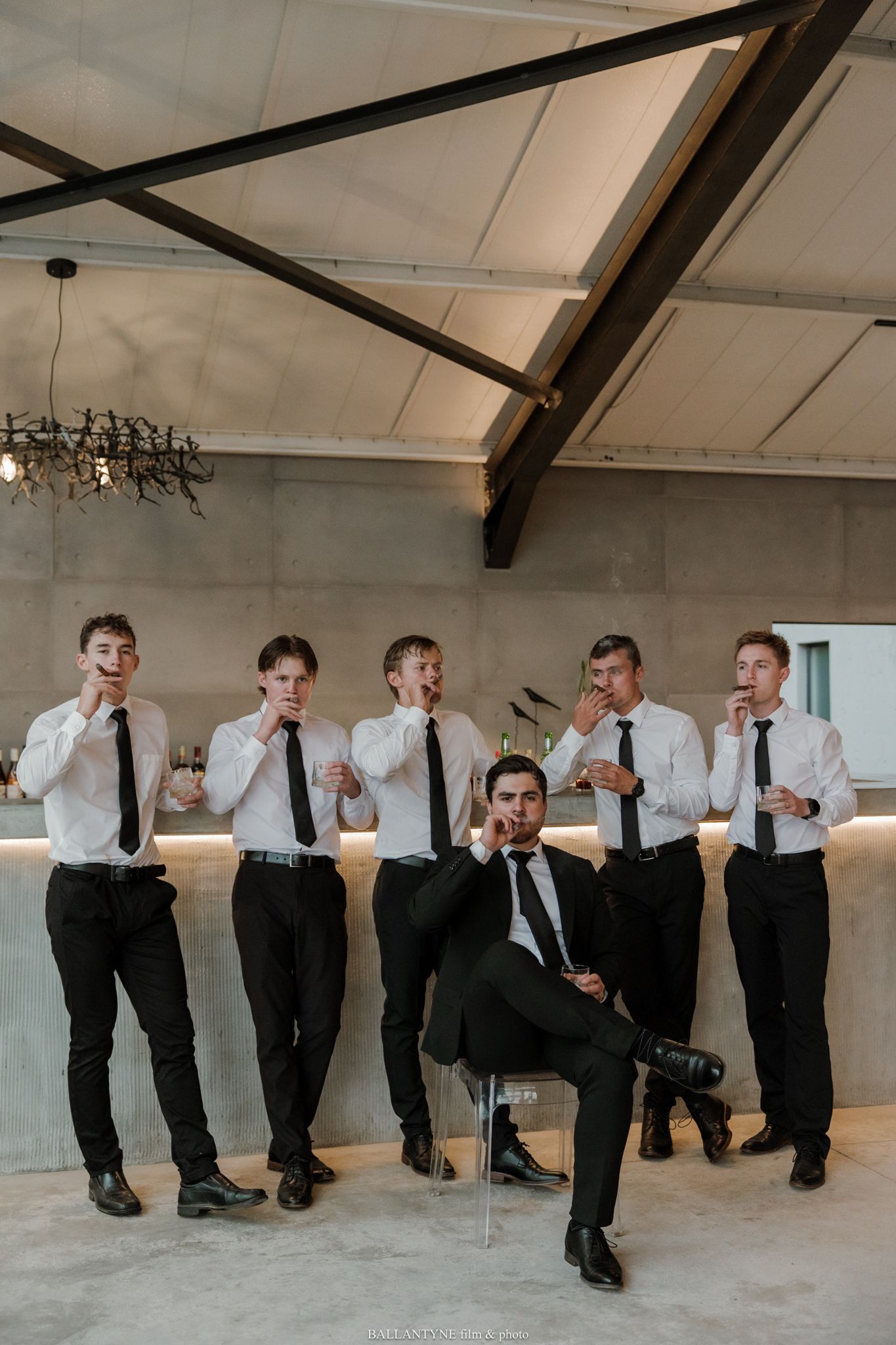Groom in black suit poses with groomsmen in white shirts and black ties at a modern venue bar.