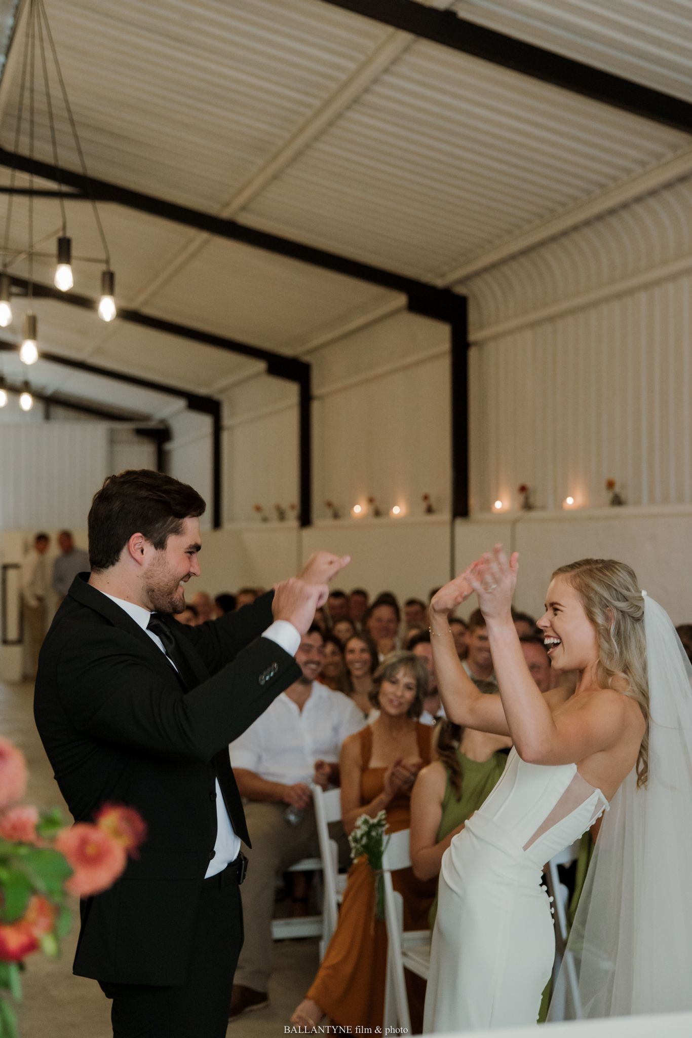 Joyful bride and groom celebrate at wedding altar in elegant barn venue with guests seated in background.