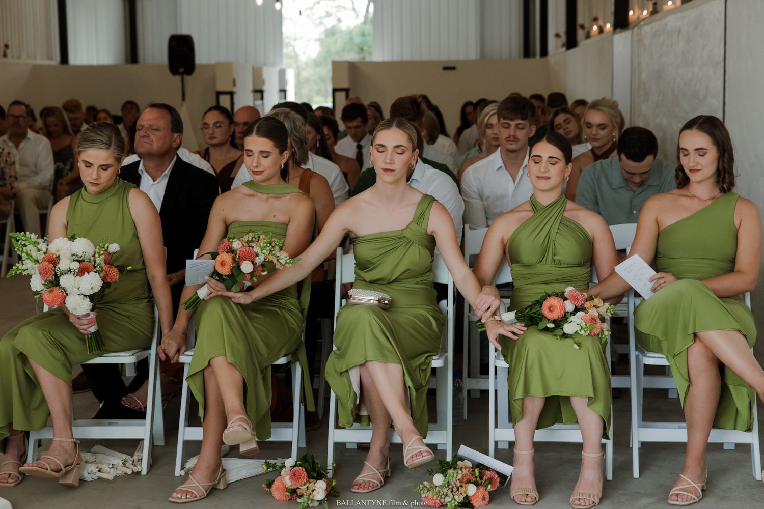 Four bridesmaids in olive green dresses holding coral bouquets seated at an outdoor wedding ceremony.
