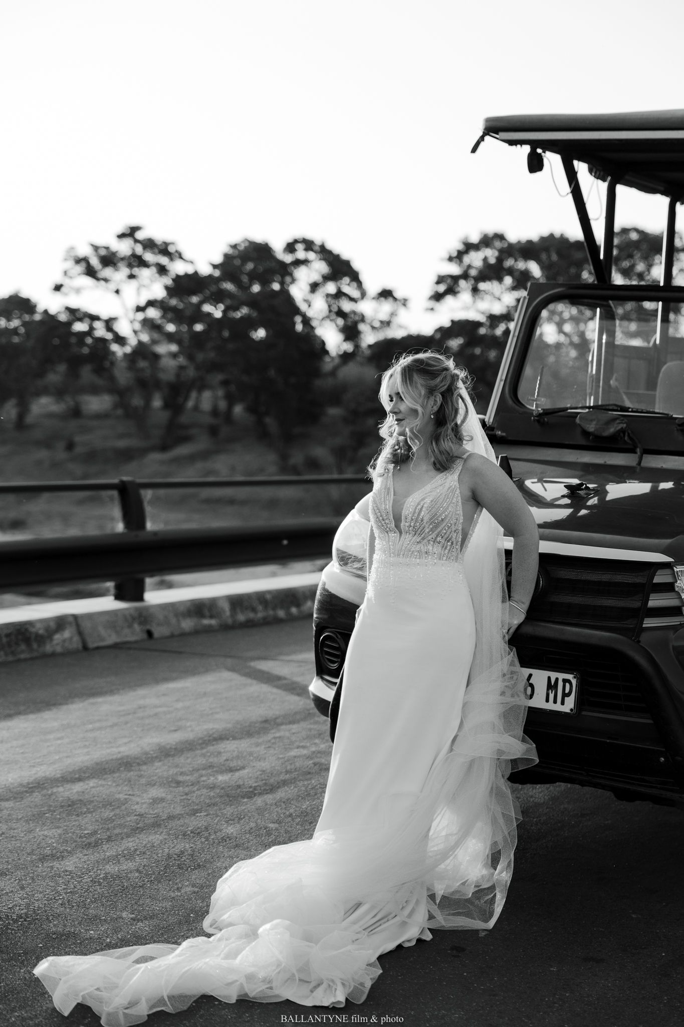 Bride in white lace dress stepping into vintage car during outdoor photoshoot.