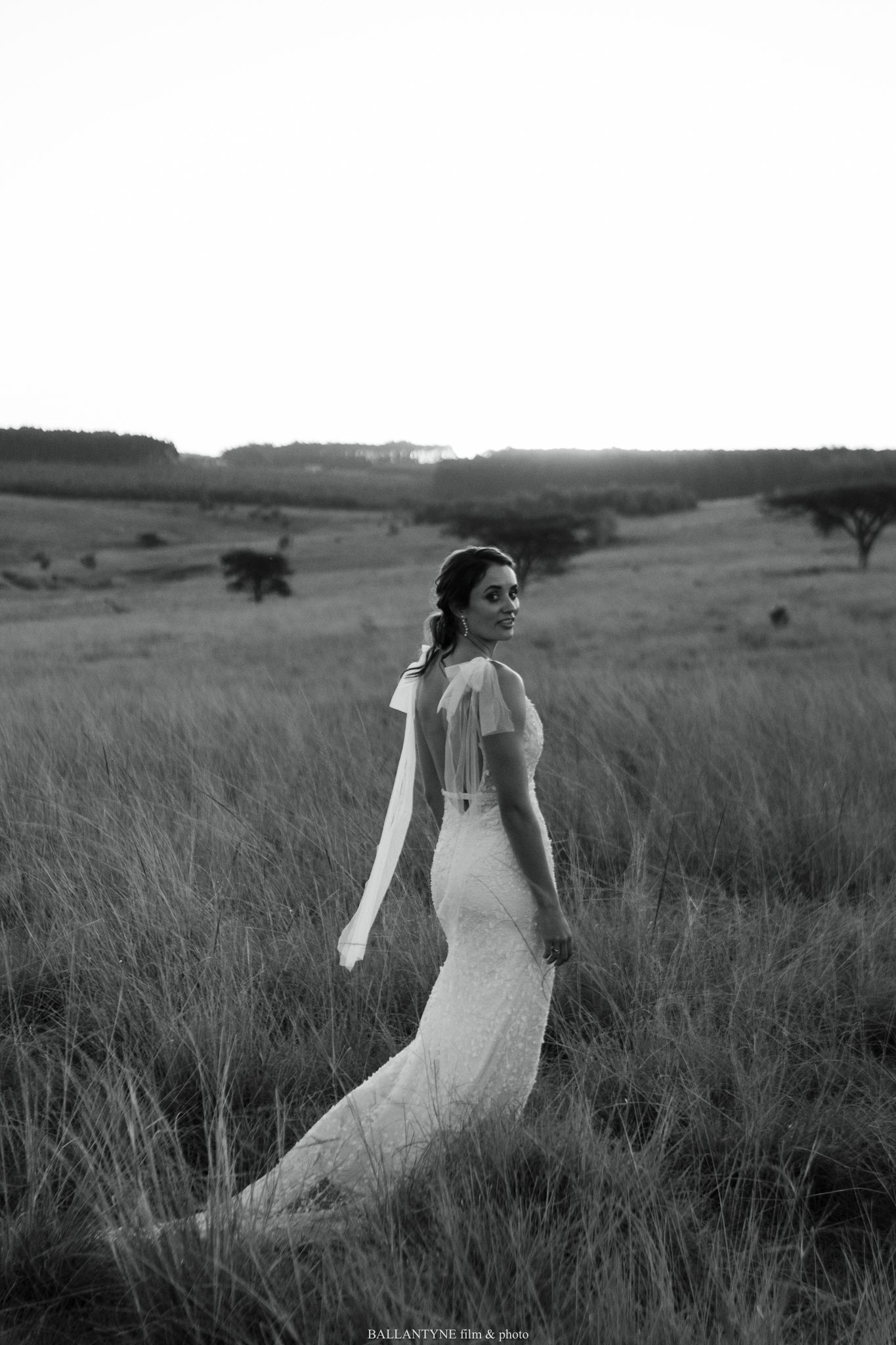 A white wedding dress flows gracefully through a serene field of tall grass at golden hour.
