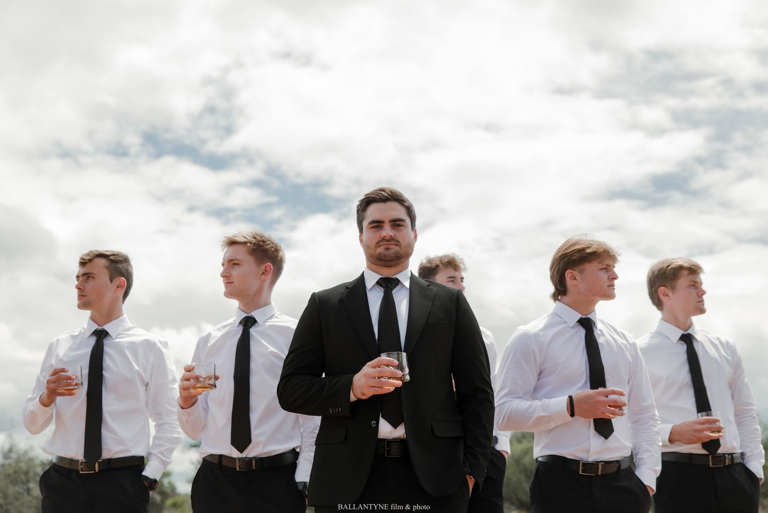 Groom in black suit stands confidently in front of groomsmen in white shirts and ties outdoors.