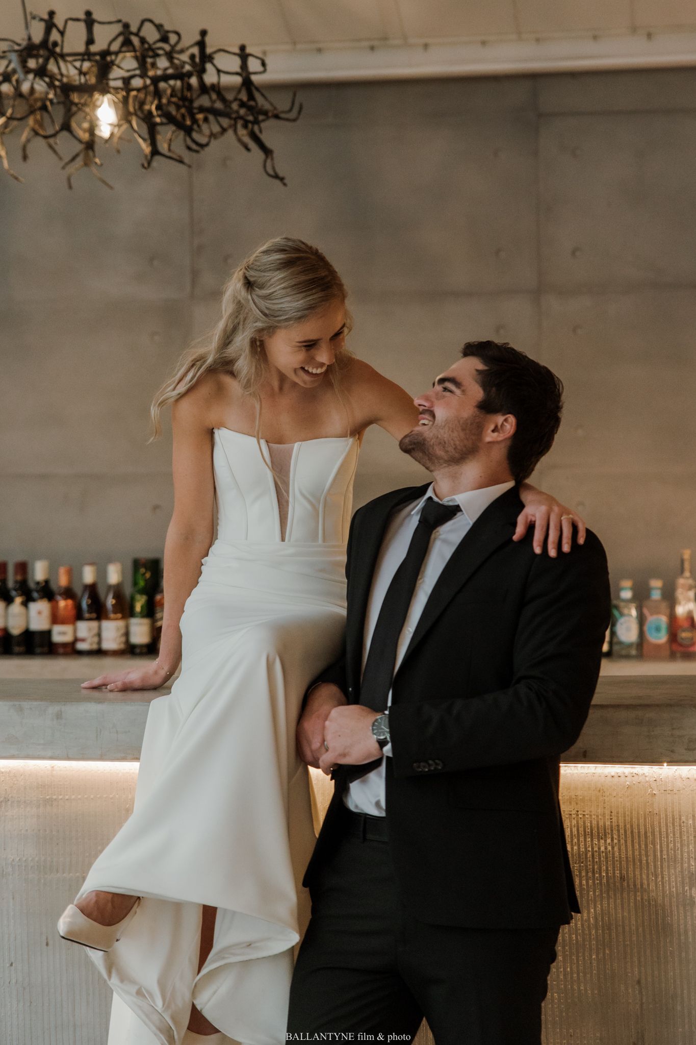 A bride in a sleek white gown sits on a bar laughing with her groom in a black suit at their wedding reception.