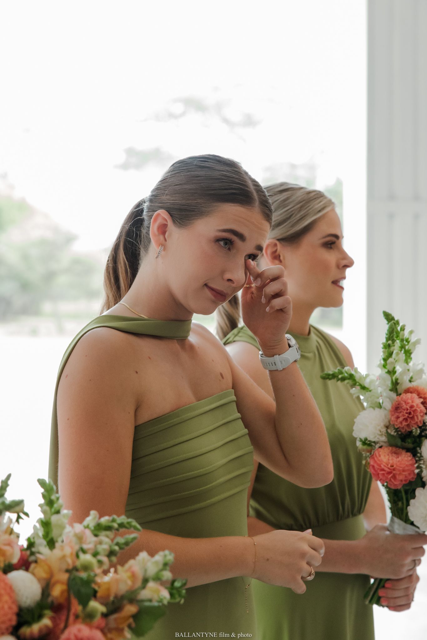 Two bridesmaids in olive green dresses holding colorful bouquets during an emotional wedding ceremony.