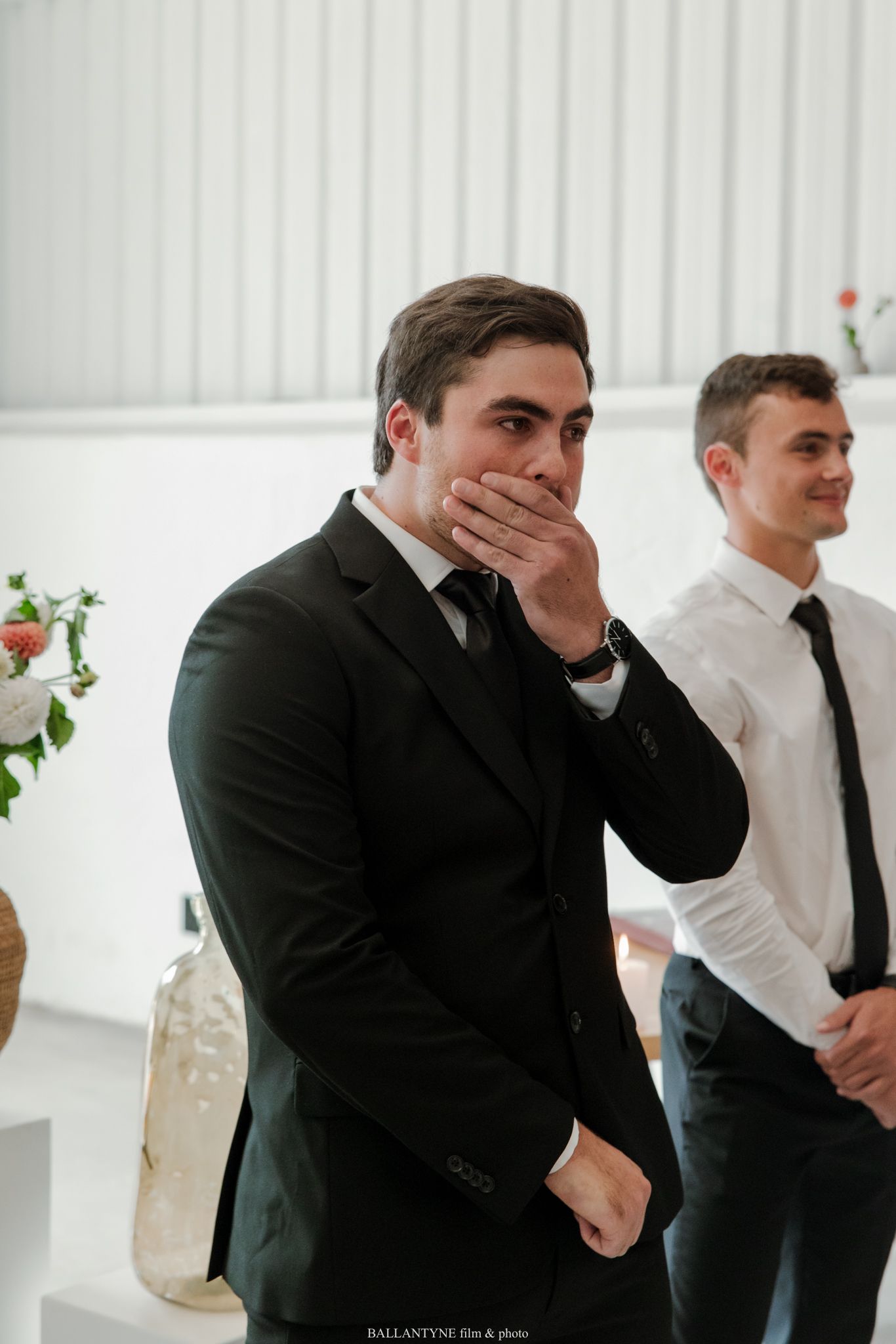Emotional groom in black suit covers mouth in awe during wedding ceremony, with groomsman in white shirt standing behind.