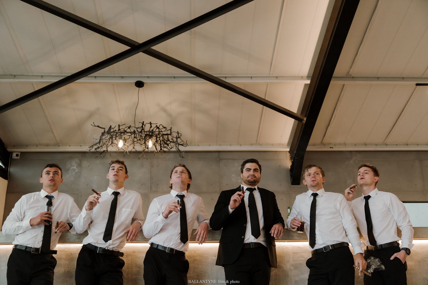 Six groomsmen in white shirts and black ties laughing and posing together at a wedding venue bar.