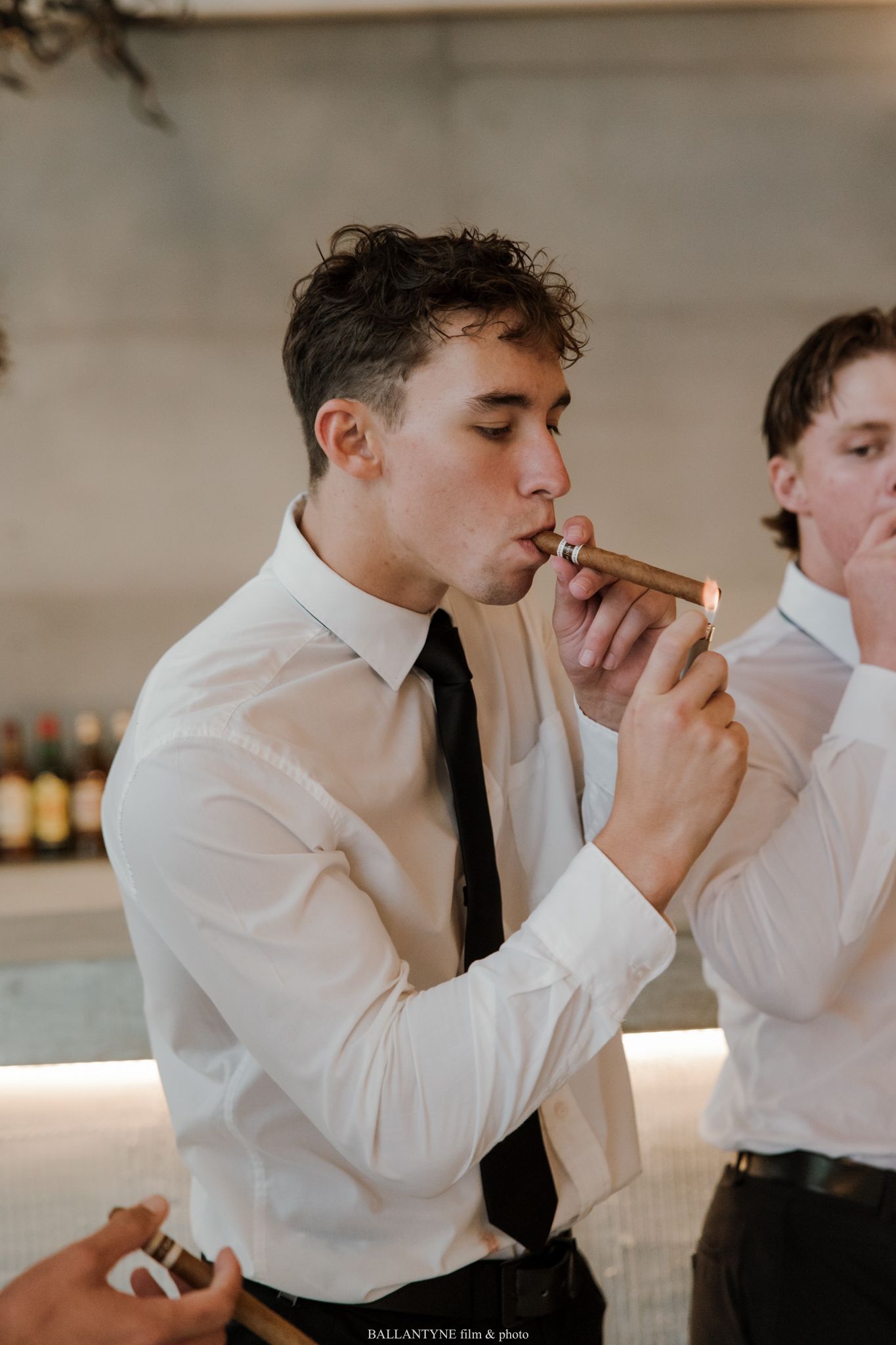 Young man in white shirt and black tie lighting a cigar at a formal event or wedding reception.