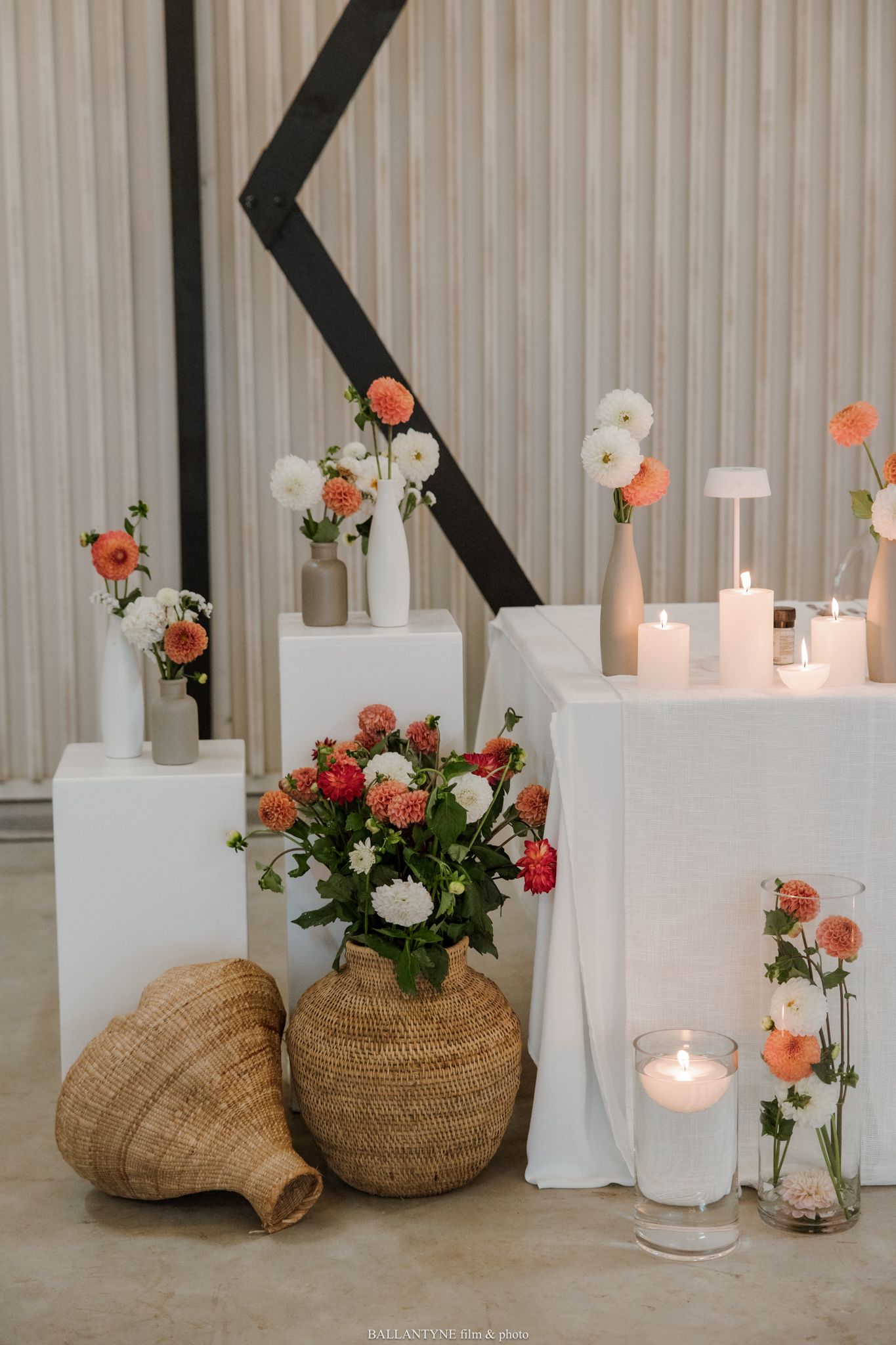 Elegant wedding decor with coral and white flowers, wicker vases, candles, and white pedestals against a neutral backdrop.
