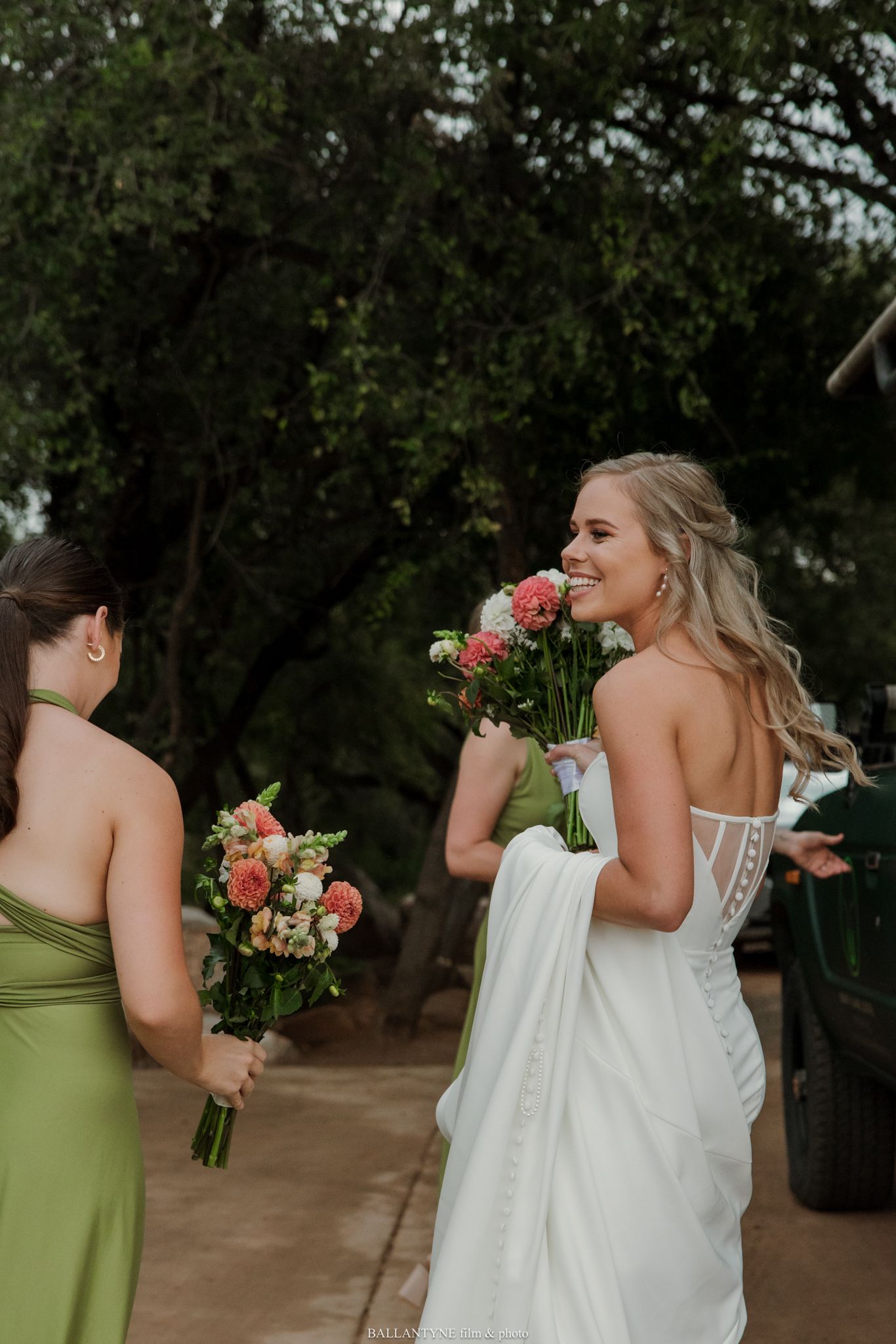 Bride in white gown smiling and holding pink bouquet with bridesmaid in green dress outdoors.