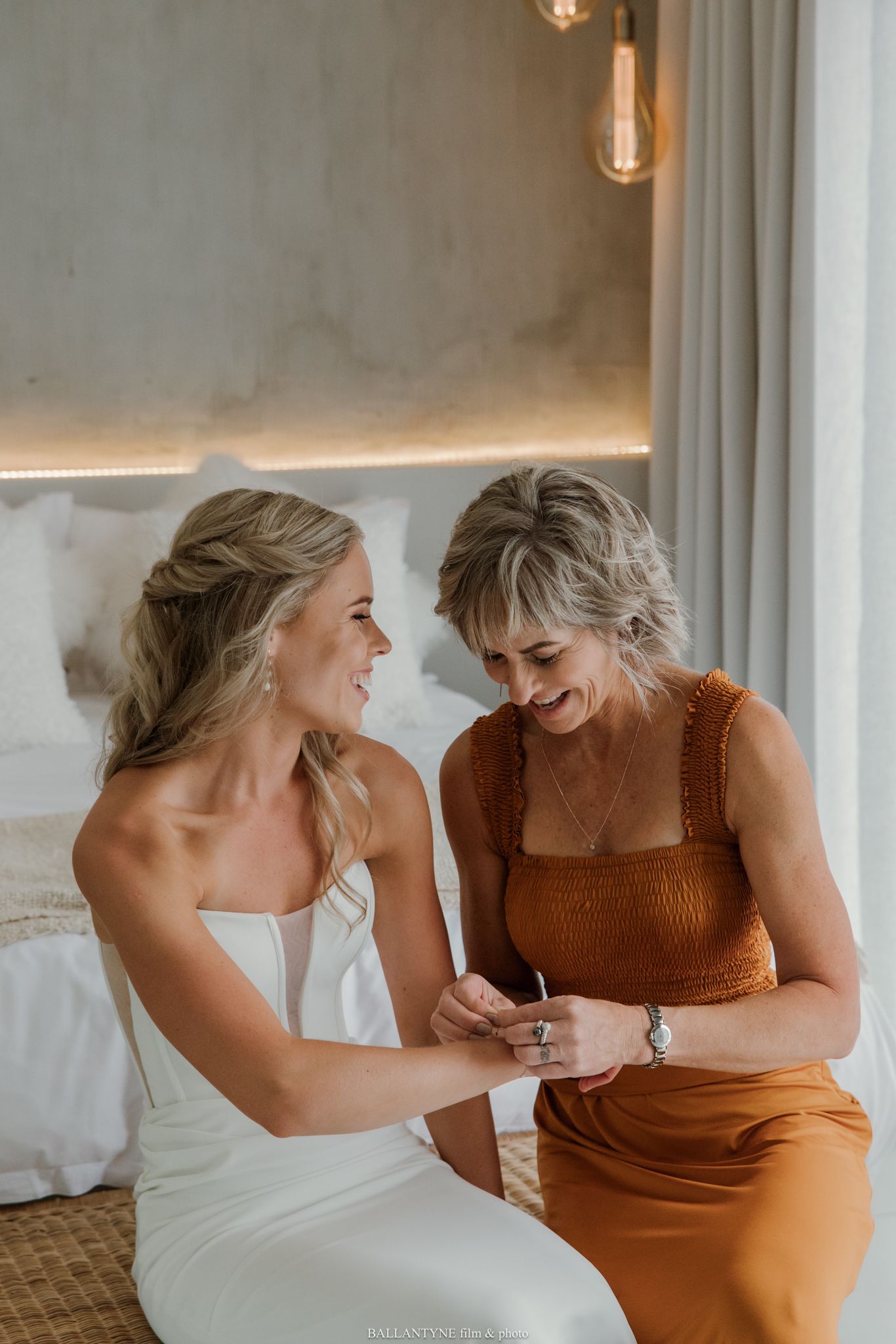 Bride in white gown sits with woman in rust dress, sharing a joyful moment while looking at jewelry.