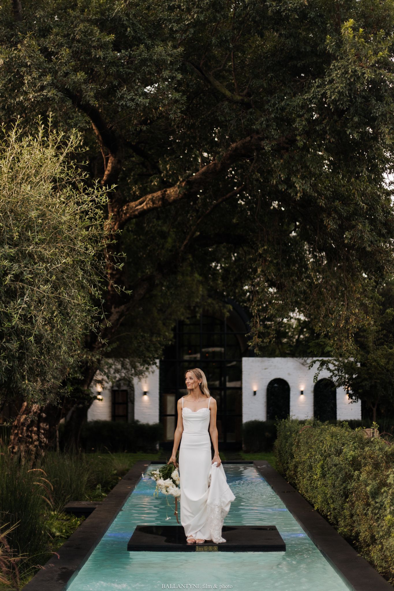 Wedding party members pose around a modern pool setting surrounded by greenery.