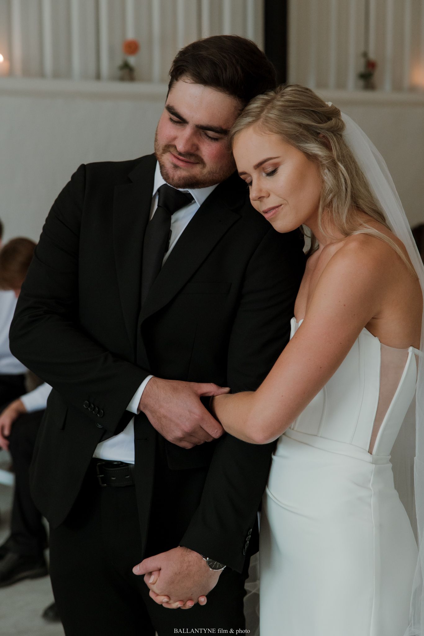 Bride and groom share an intimate moment during wedding ceremony, groom in black suit, bride in white gown with veil.