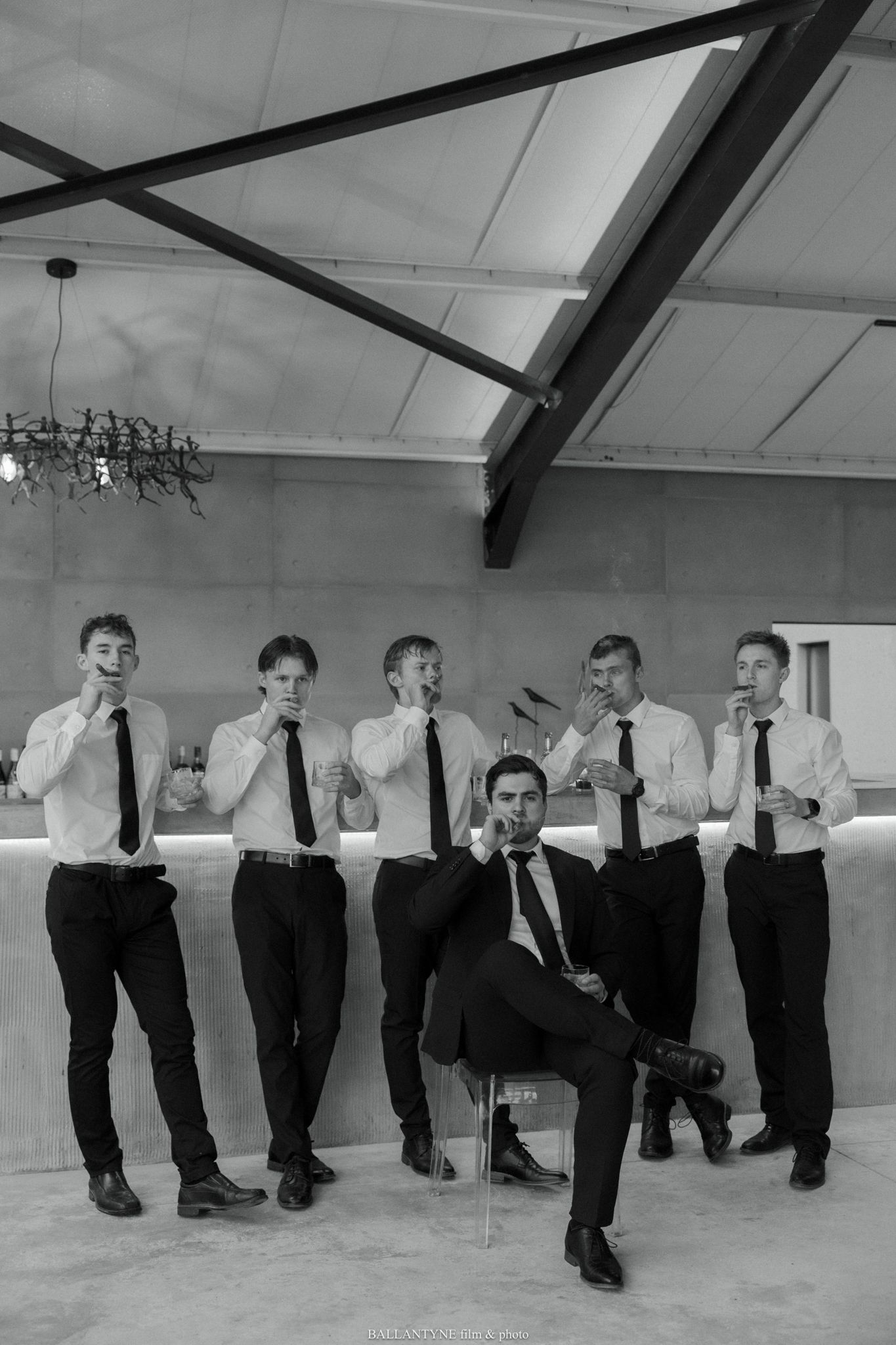 Groom sits confidently in a chair while five groomsmen in white shirts and ties pose behind him in a modern venue.