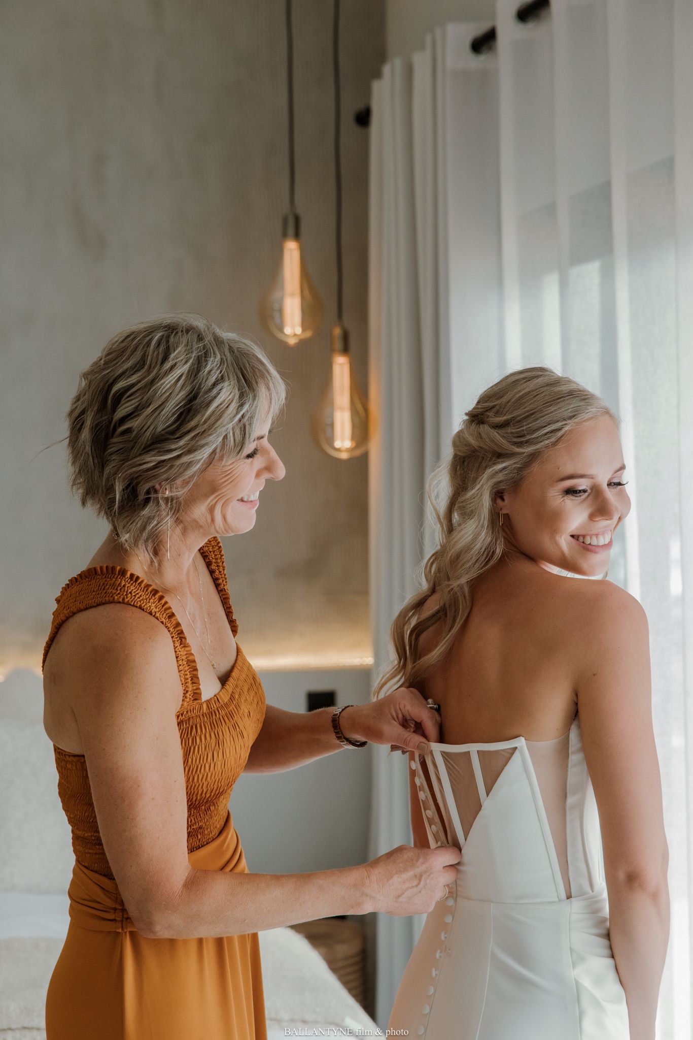 A mother helps her smiling bride daughter button up her white wedding dress in a softly lit bridal suite.