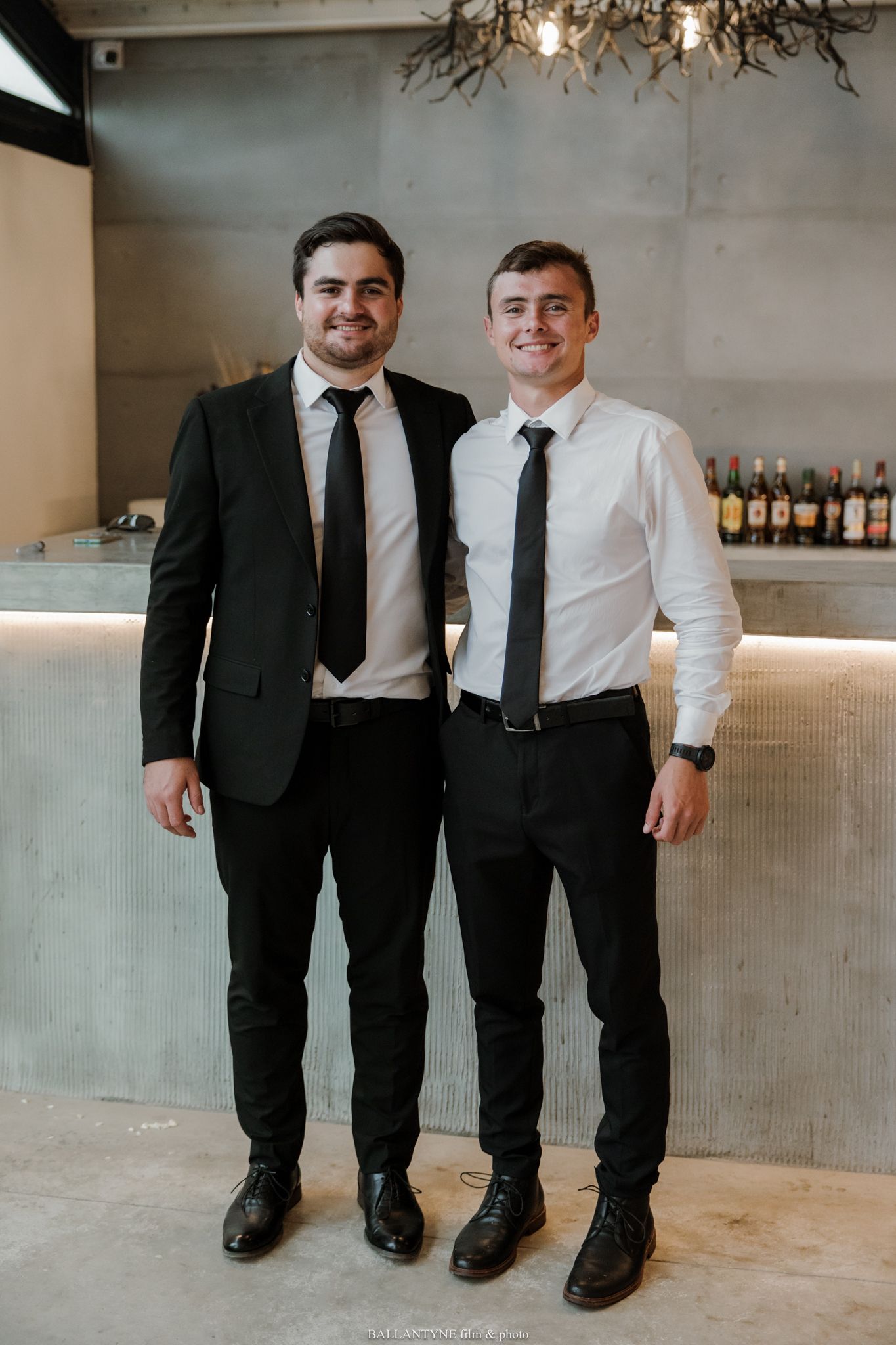 Two young men in black suits and ties smiling together in front of a bar backdrop.
