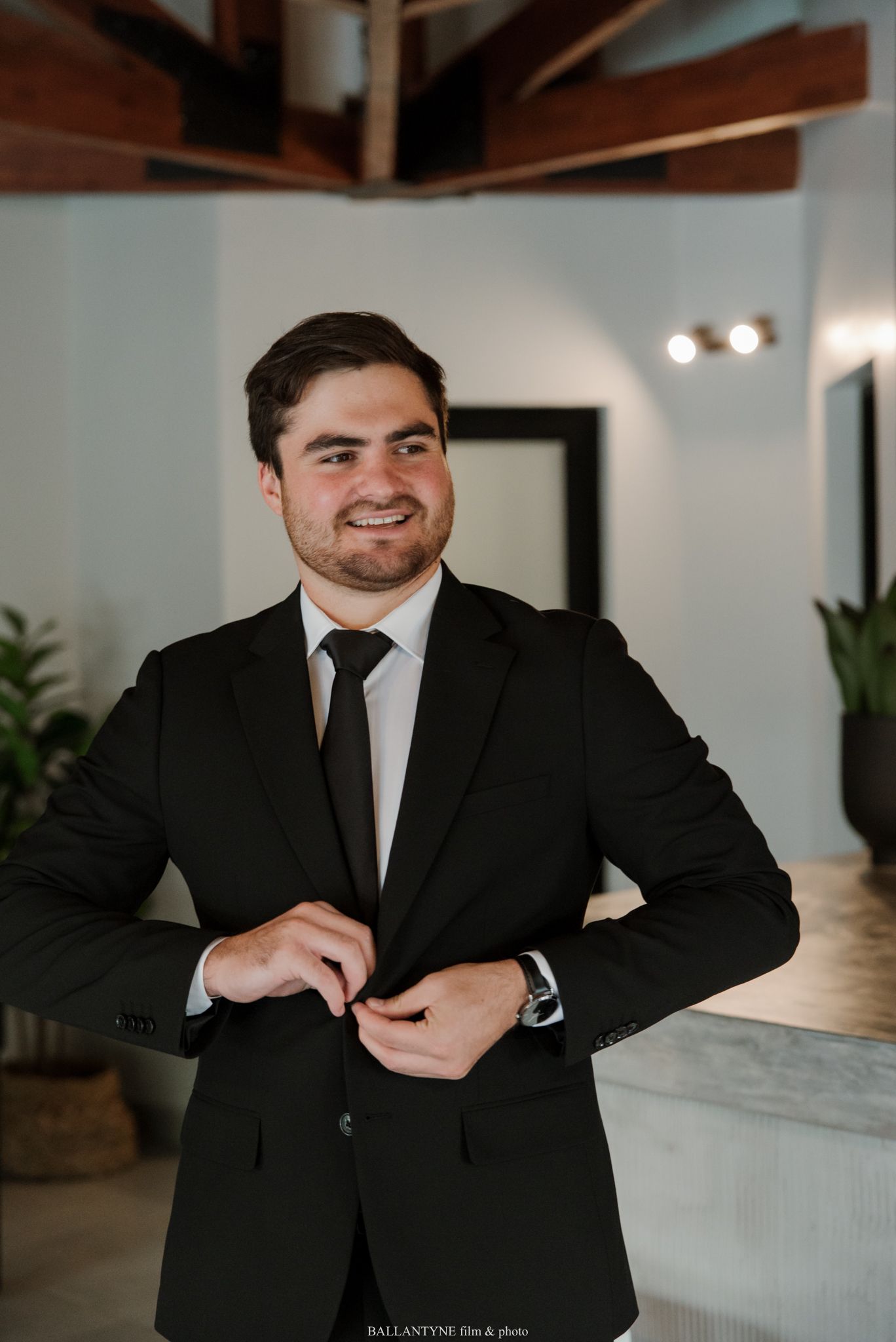 Smiling groom in black suit and tie buttoning jacket indoors before wedding ceremony.