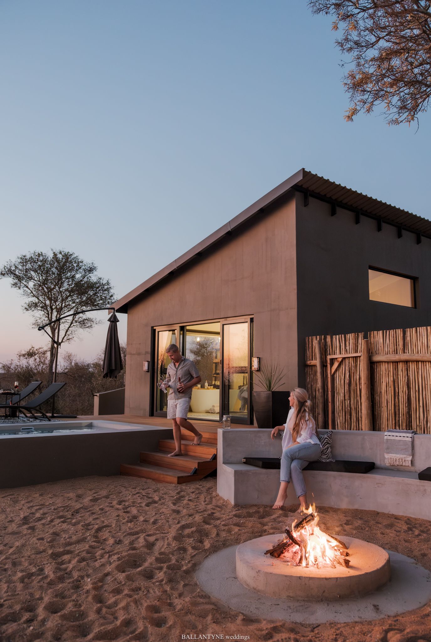 Couple relaxing by a fire pit at dusk outside a modern safari lodge with a plunge pool in the background.