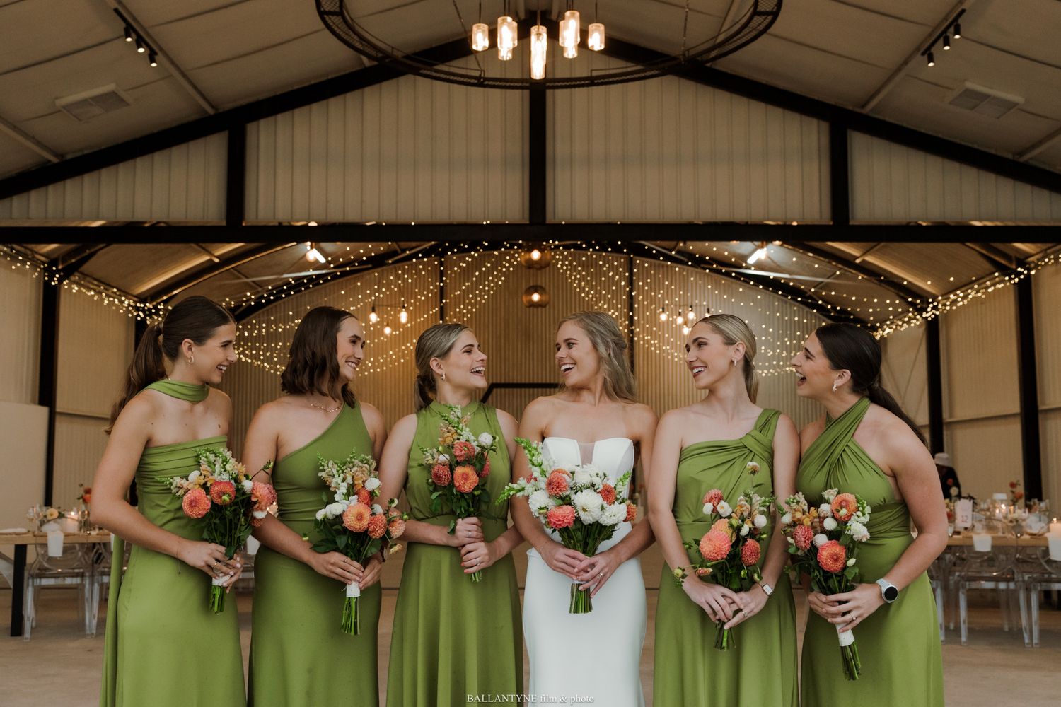 Bride in white gown with bridesmaids in green dresses holding coral bouquets inside a rustic barn wedding venue.