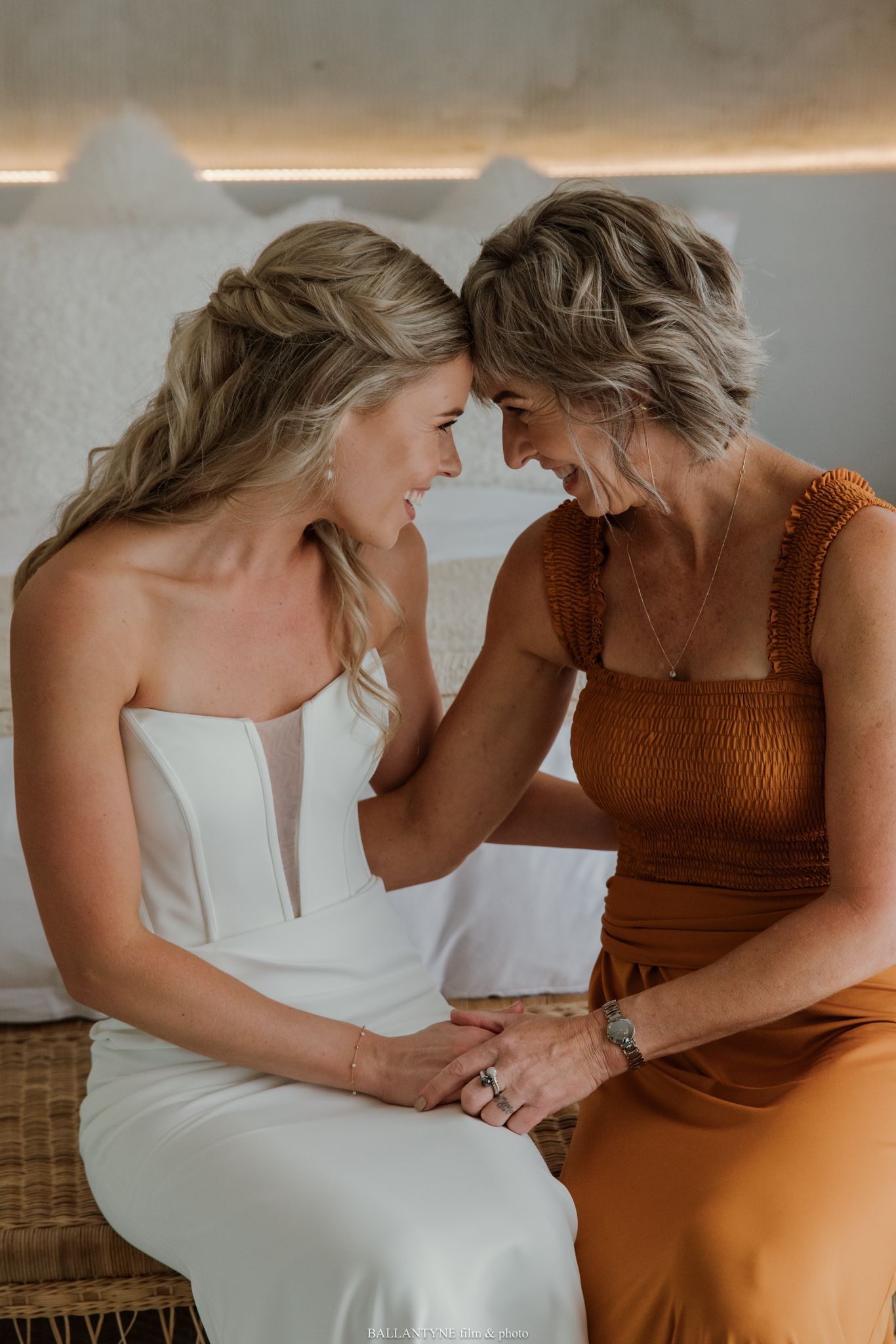 A bride in a white strapless gown shares a tender moment with an older woman in a rust dress, foreheads touching.