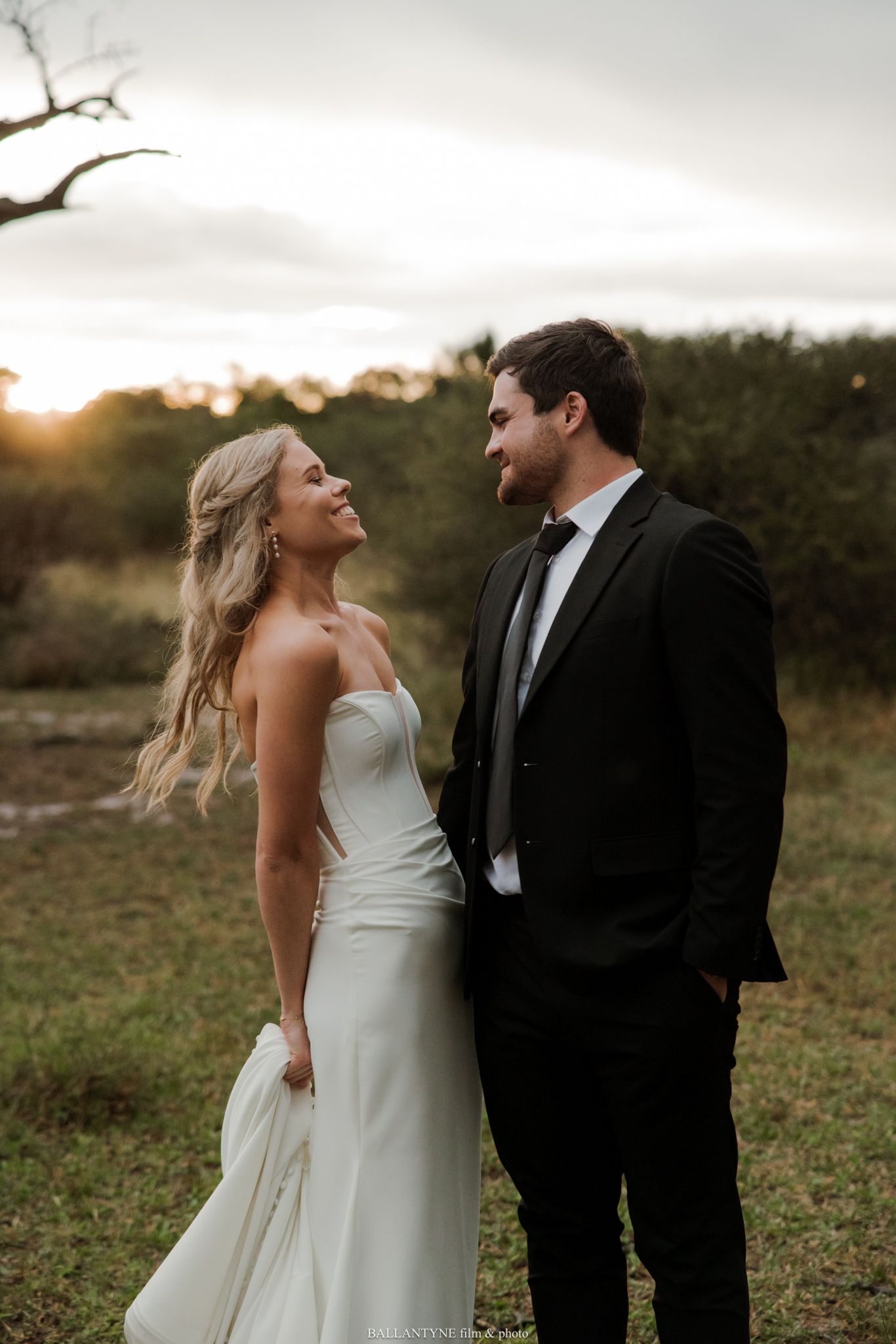 A bride in a white gown and groom in a black suit share a romantic moment outdoors at golden hour.