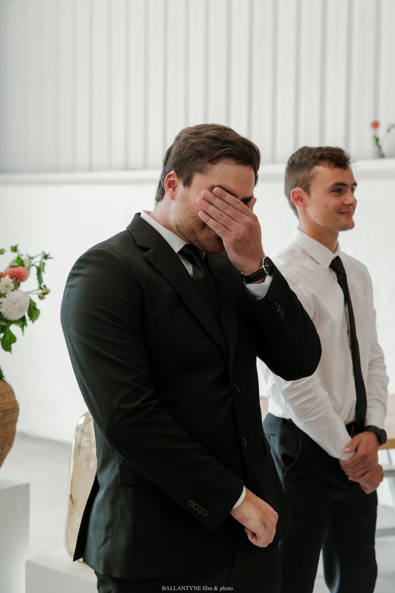 Emotional groom covering his face during wedding ceremony, with smiling groomsman in white shirt standing behind him.