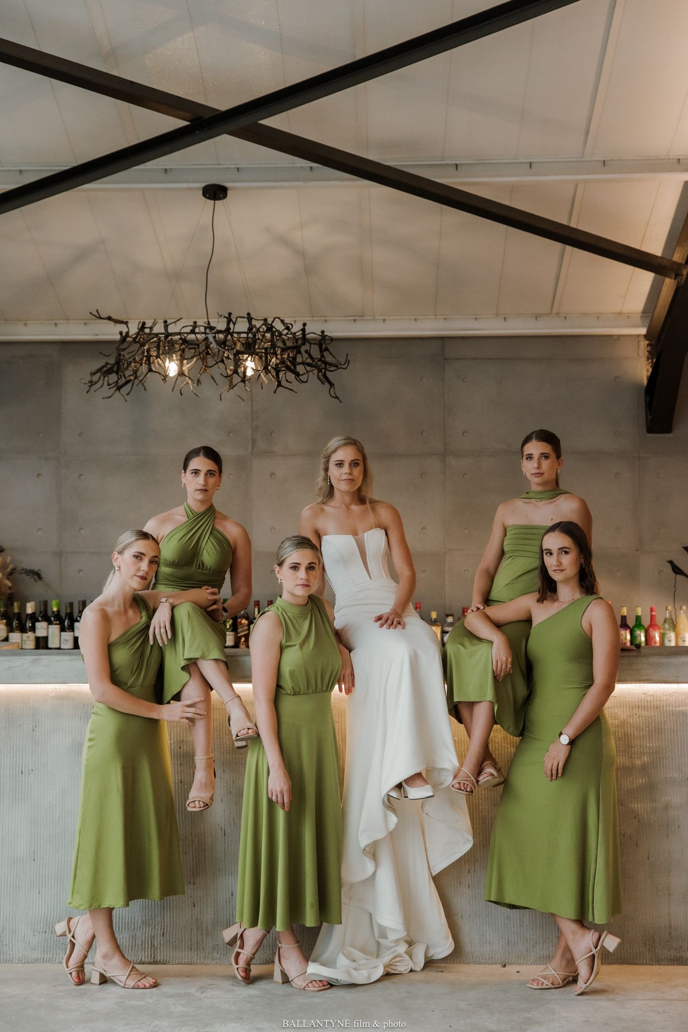 Bride in white gown poses with five bridesmaids in olive green dresses inside an elegant venue.