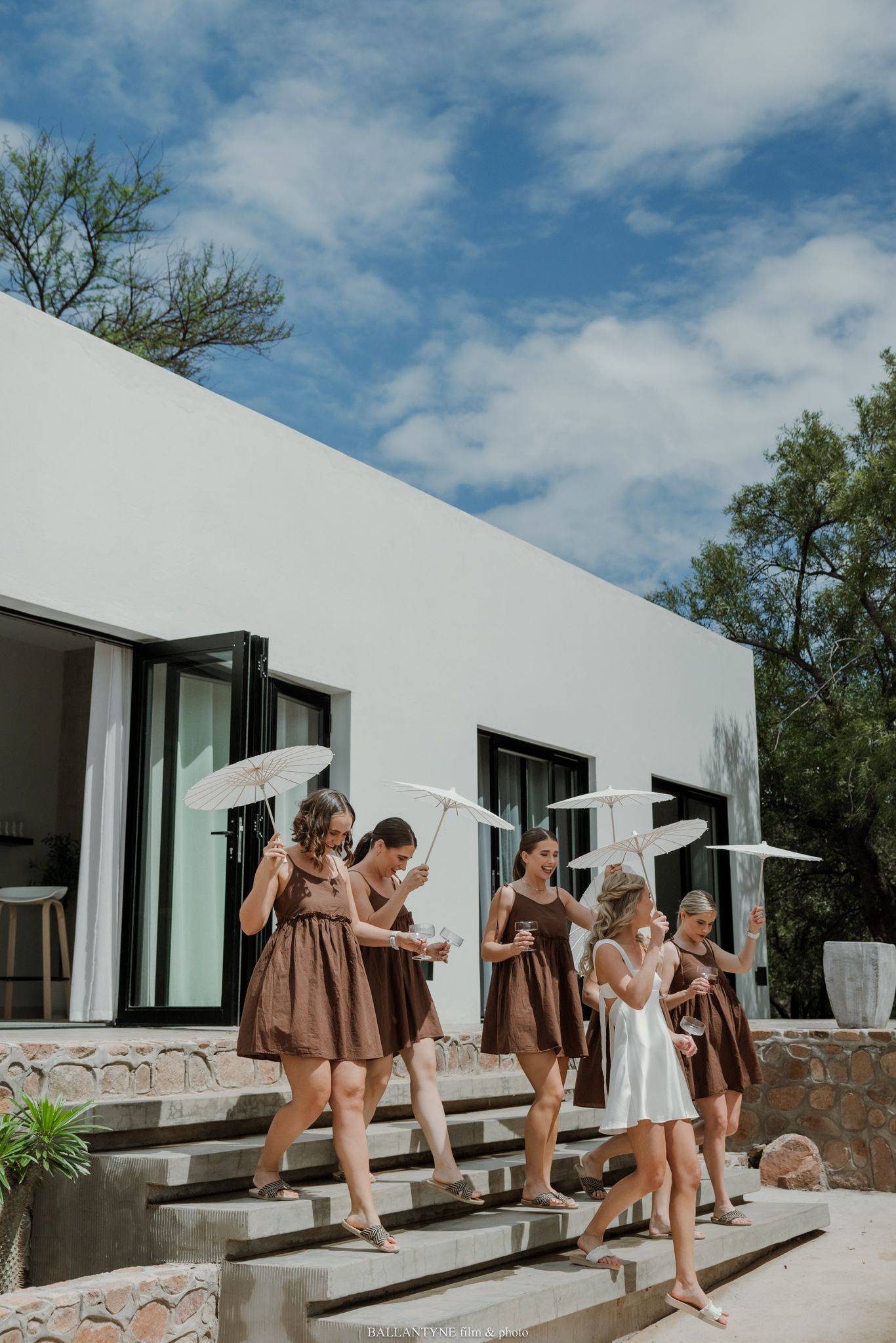 Bridesmaids in brown dresses holding white parasols walk down stone steps outside a modern white building.